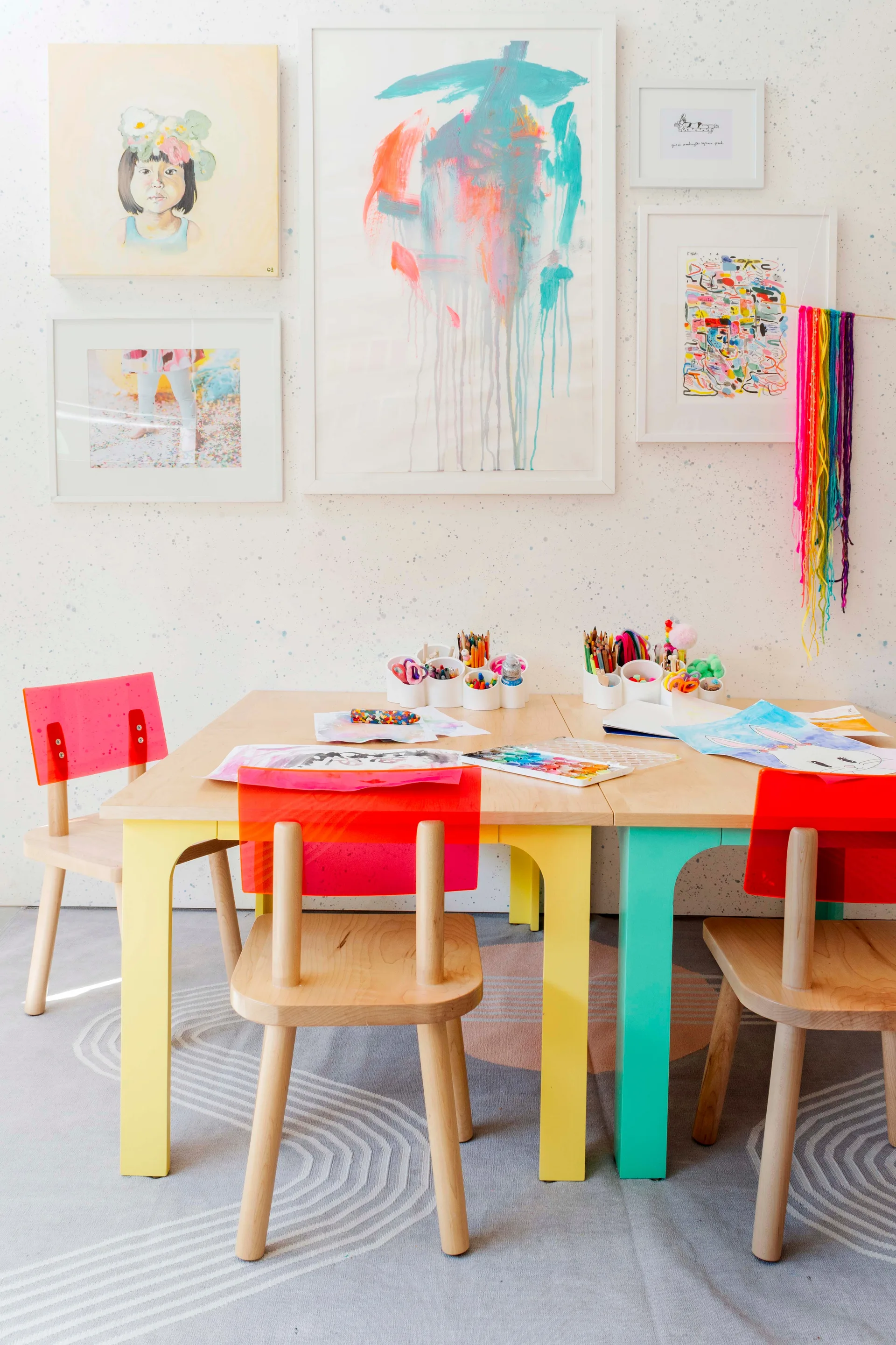 A wooden children's craft table is surrounded by wood chairs with bright pink and red acrylic backs beneath a gallery wall of framed art.