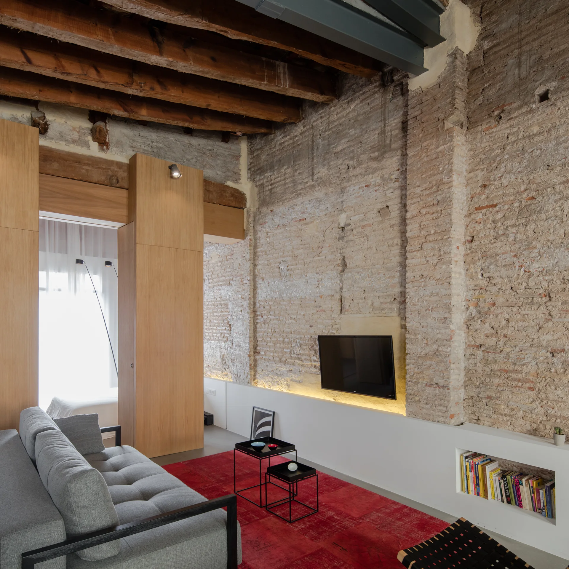 A modern living room featuring a grey sofa, a bold red area rug, and an exposed brick wall with wood ceiling beams overhead.