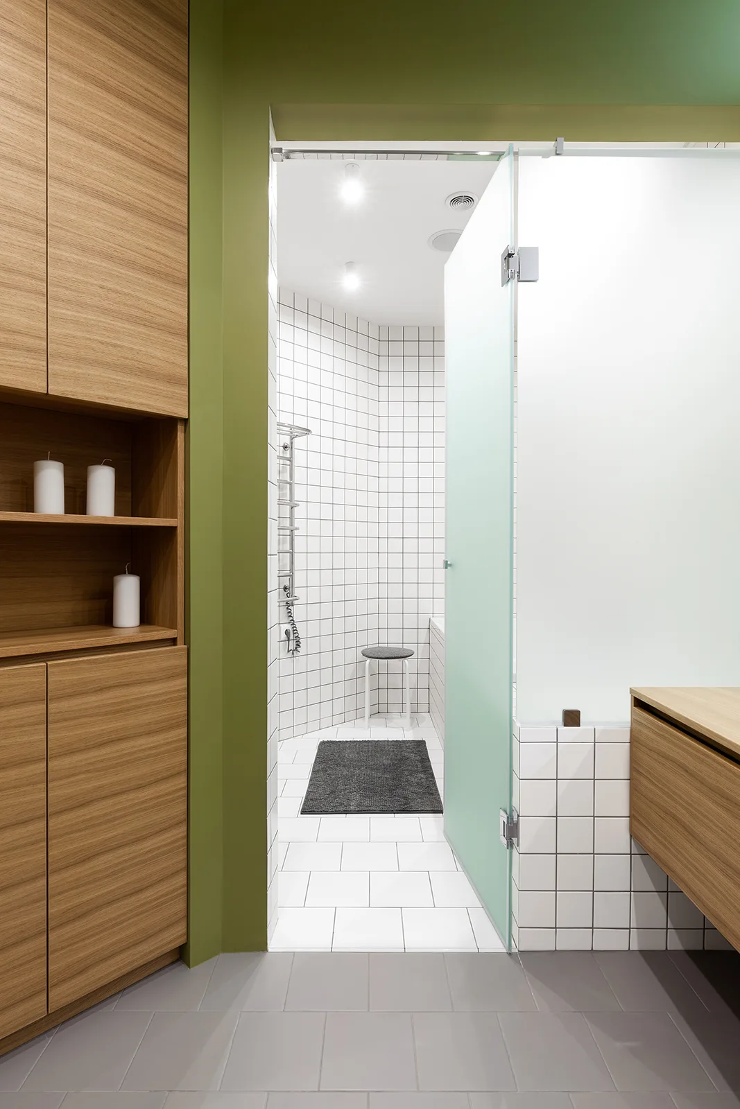 A modern bathroom entryway framed in olive green paint features built-in wood cabinetry and leads to a white grid-tiled shower area.