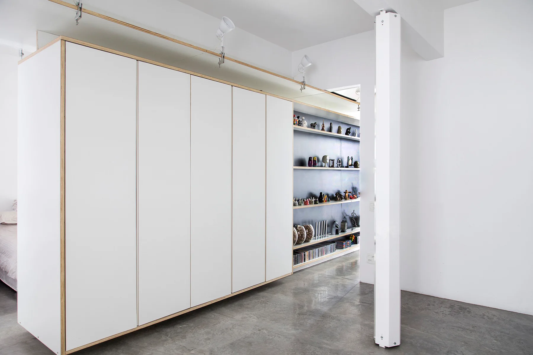 A large floor-to-ceiling white wardrobe with exposed plywood edges stands on a polished concrete floor next to integrated open shelving.