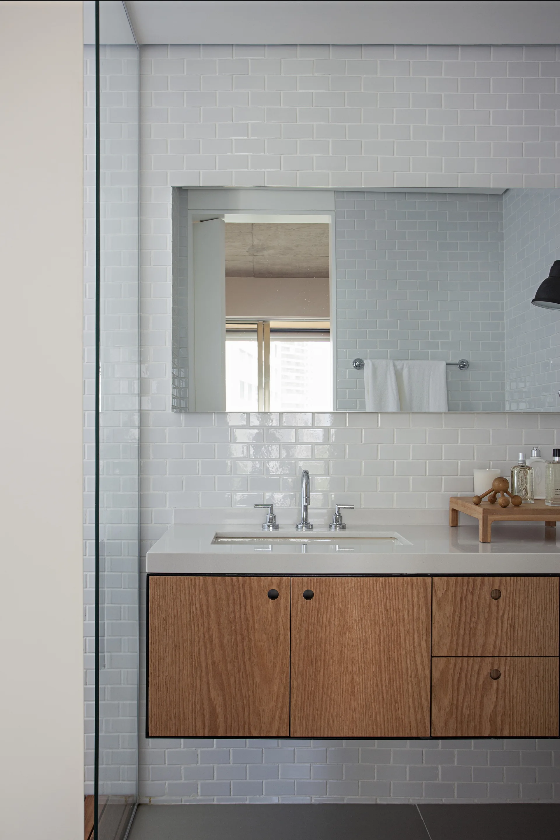 A modern bathroom features a wood floating vanity with a white countertop and sink below a glossy white subway tile wall and a large mirror.