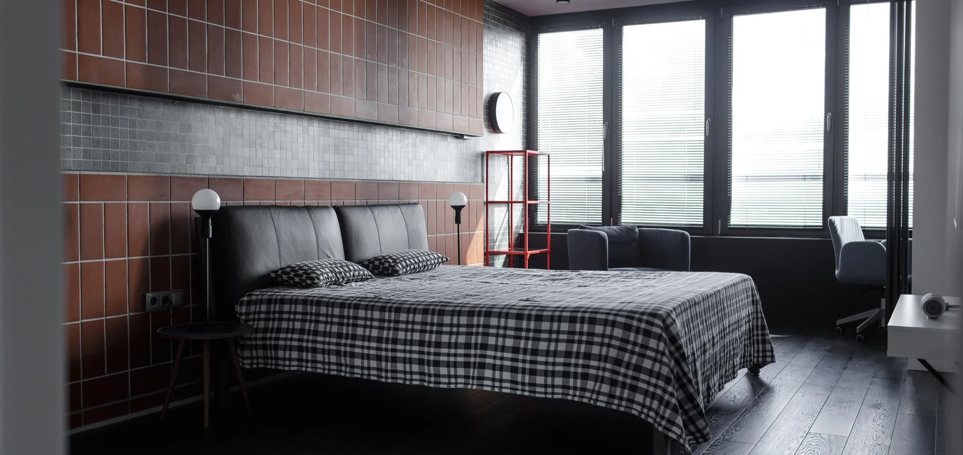 A modern bedroom features a bed with checked sheets, a dark headboard, and a large terracotta tiled accent wall next to window blinds.