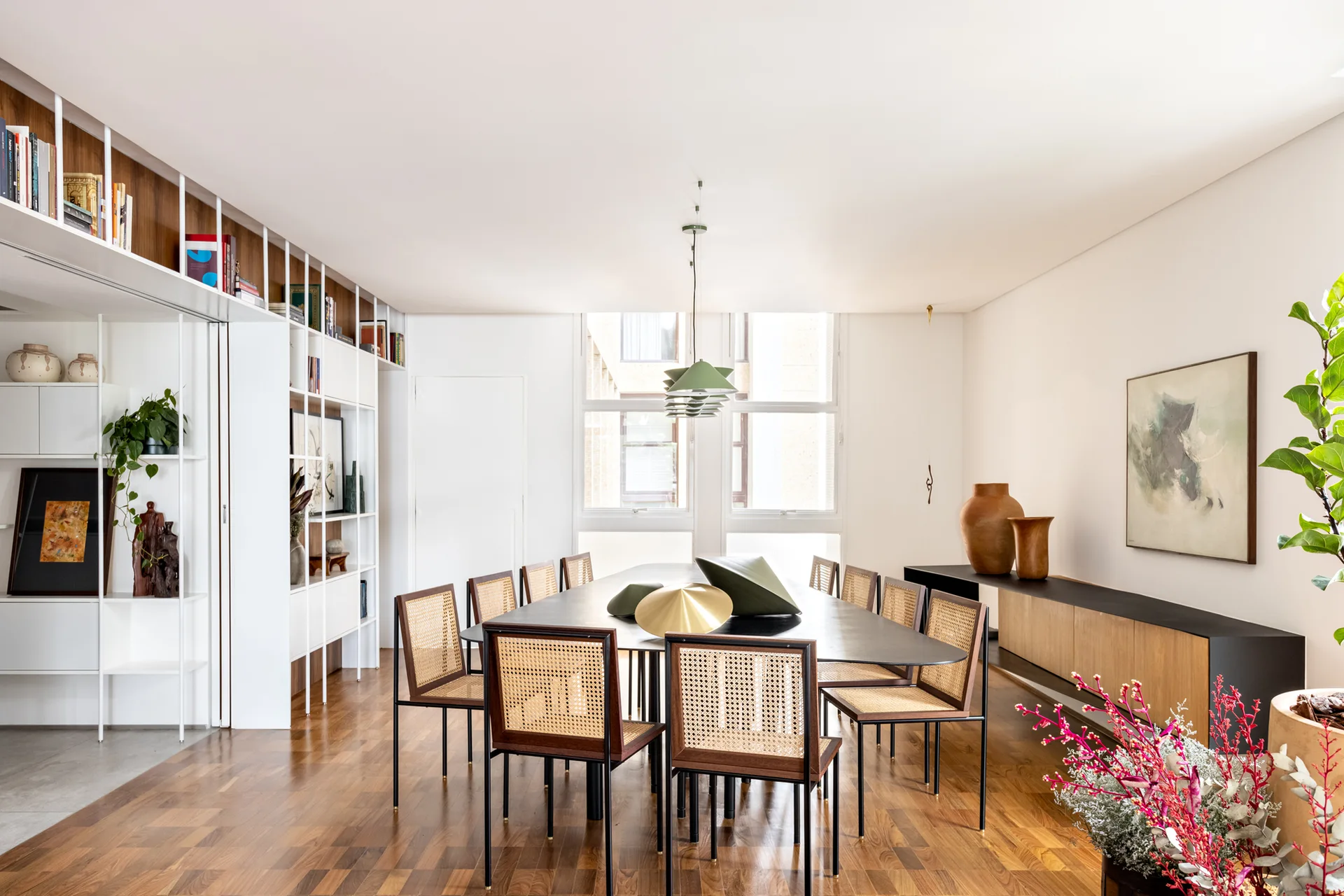 A contemporary dining room features a black oval table surrounded by cane-back chairs, set against white built-in shelving and warm wood floors.