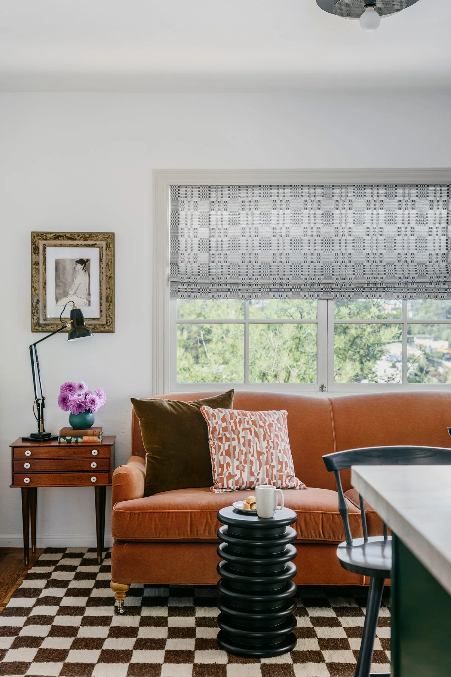 A rust-colored velvet sofa sits on a brown and white checkered rug in front of a window with a patterned Roman shade.