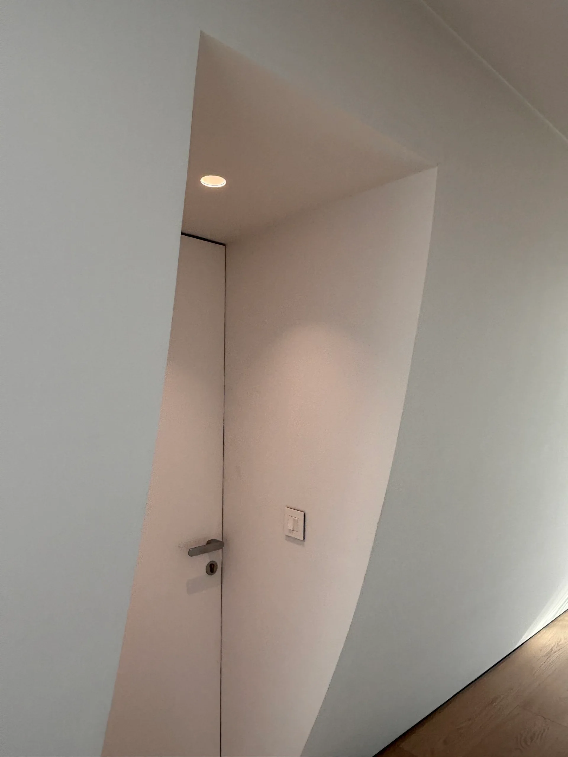 A minimalist white flush-mounted door is set within a recessed alcove featuring a single overhead downlight and a simple metal handle.