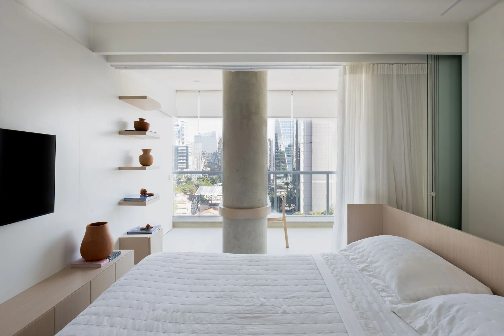 A minimalist white bedroom featuring a central concrete column, light wood floating shelves with terracotta vases, and a view of the city.