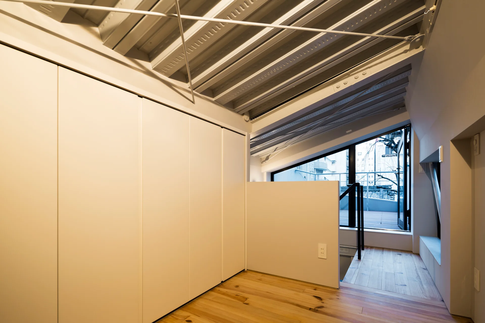 A minimalist interior loft features light wood floors, a full wall of white flat-panel storage cabinets, and an exposed white corrugated metal ceiling.