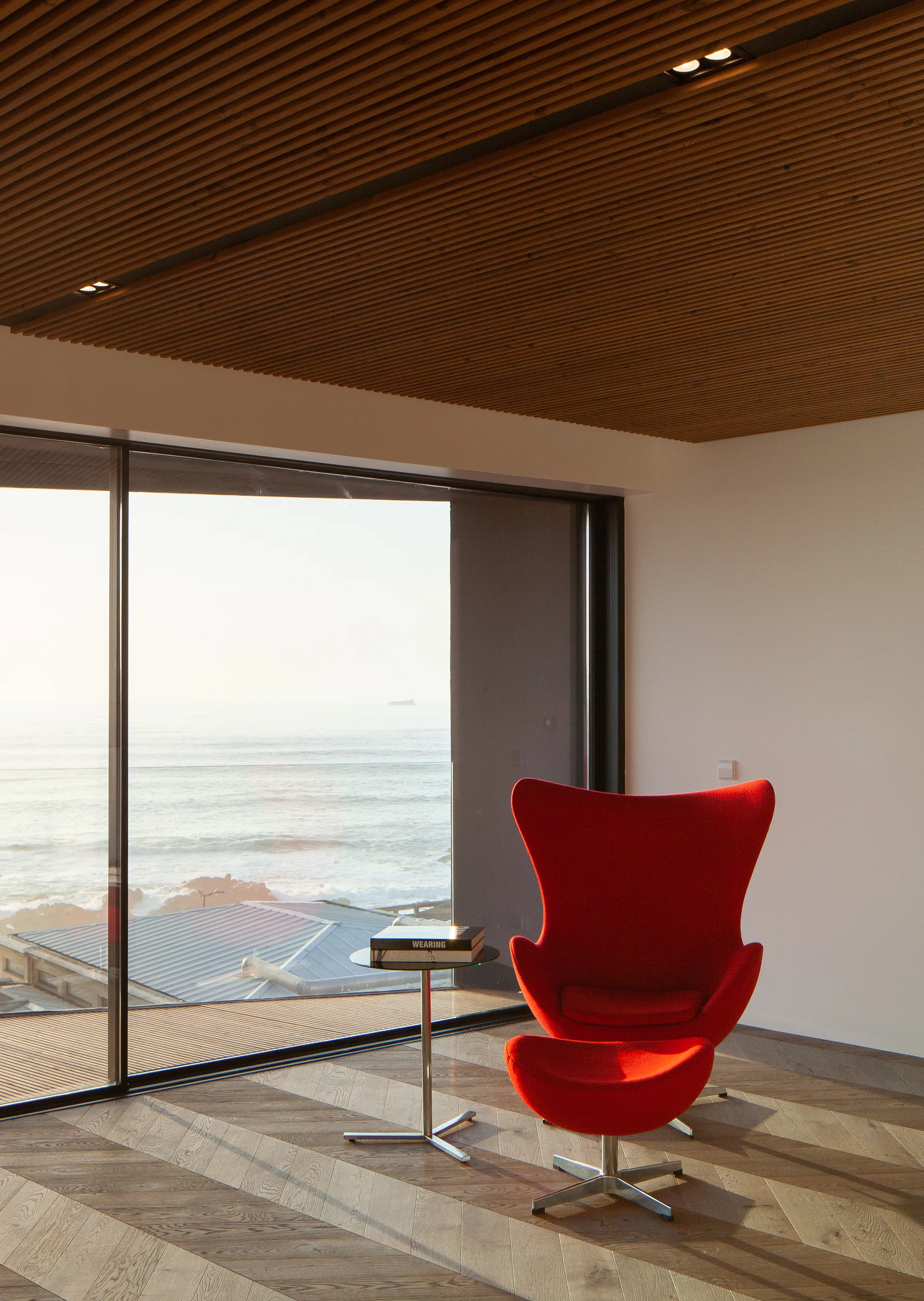 A minimalist room features a bright red lounge chair and matching ottoman on herringbone wood floors near a large window overlooking the ocean.