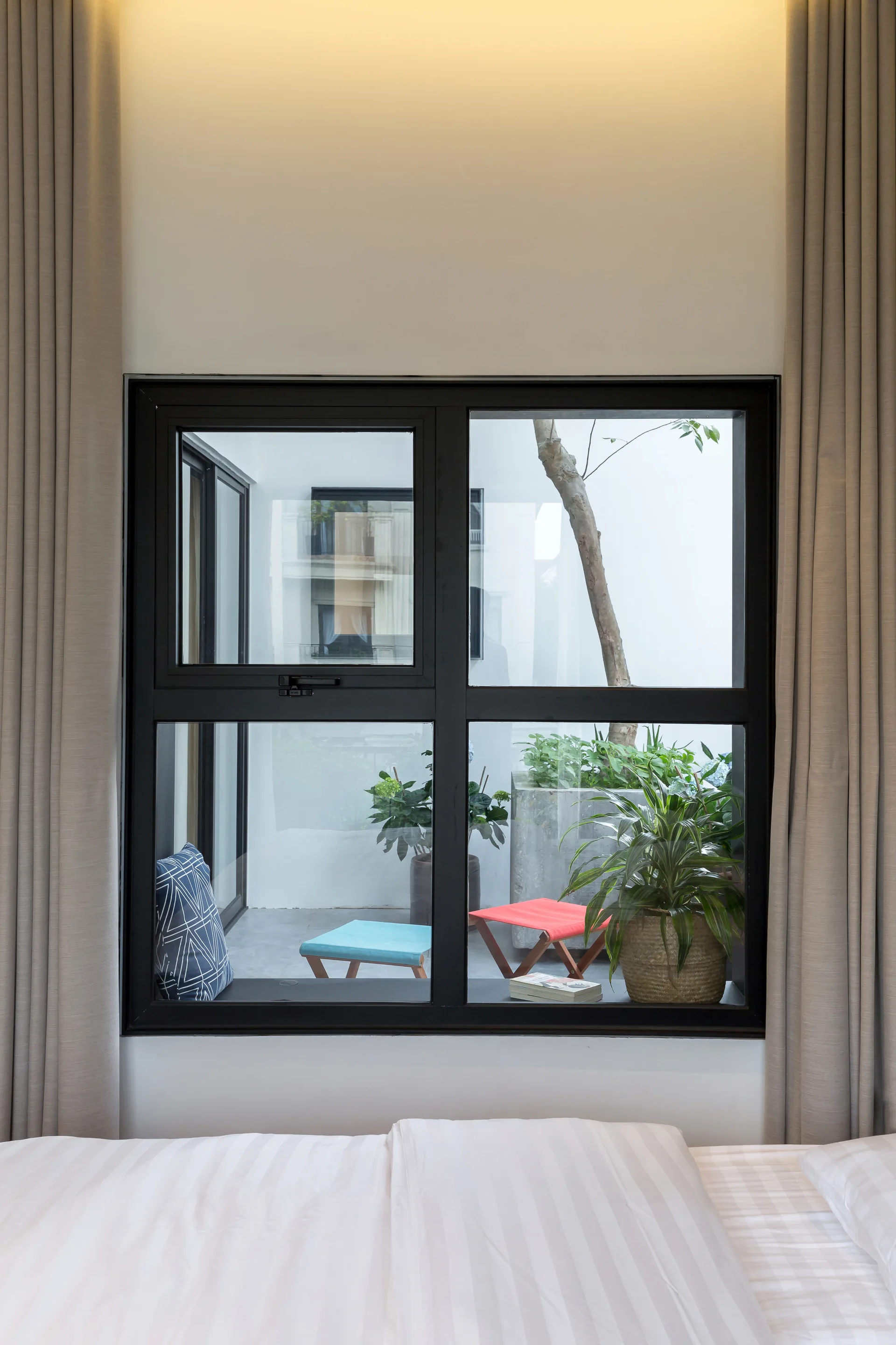 View from a bed looking through a black-framed window onto a courtyard with plants and folding stools.