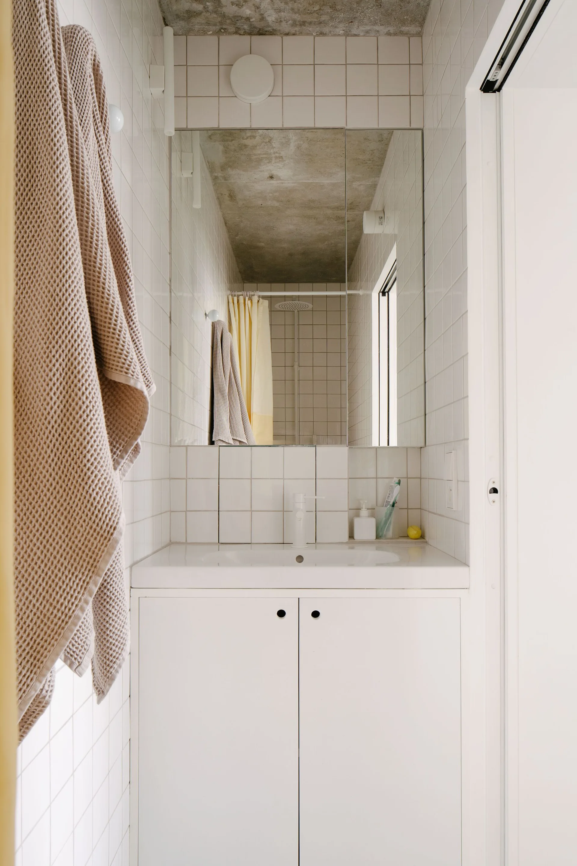 A minimalist bathroom features a white vanity, square-tiled walls, and an unfinished concrete ceiling.