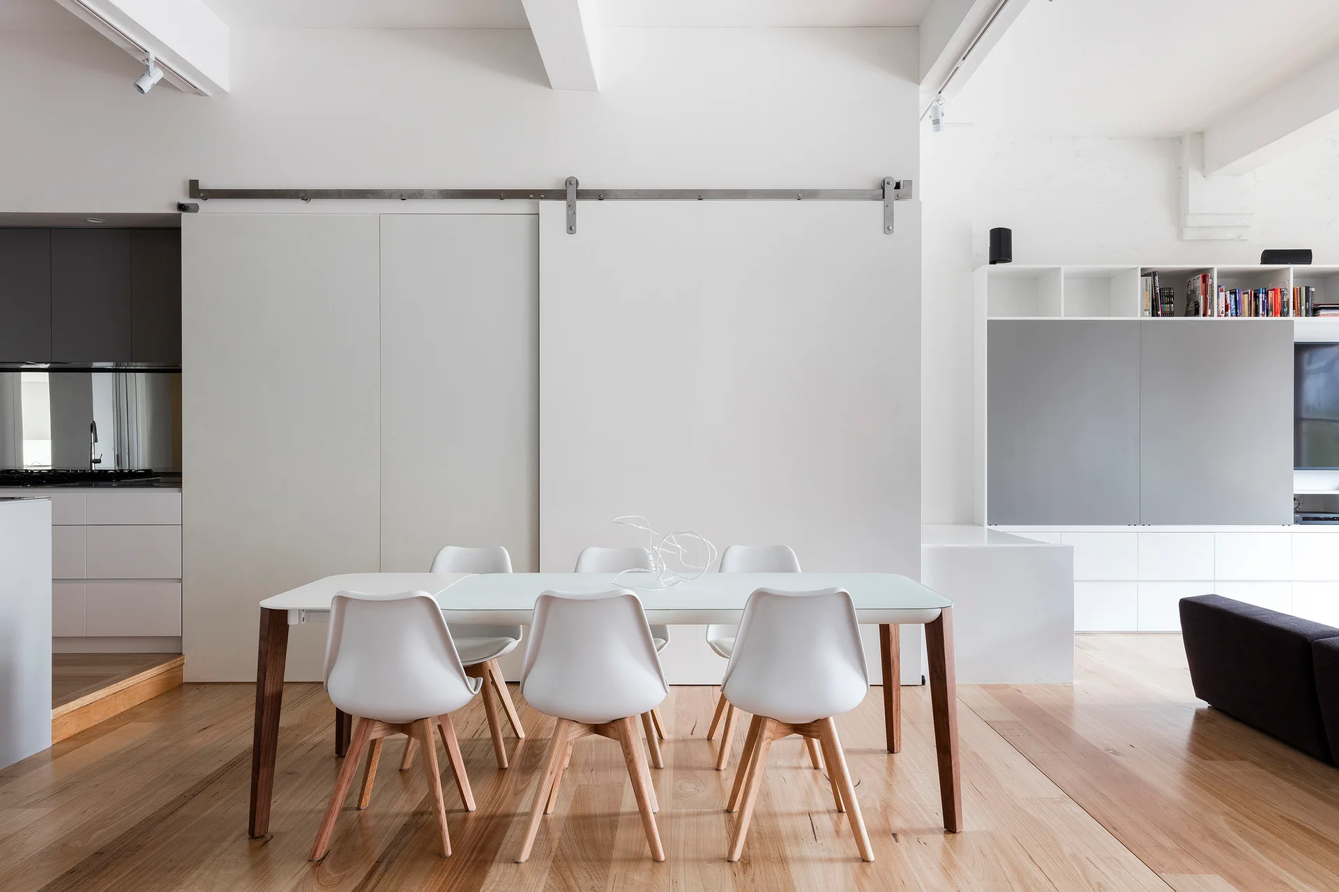 A minimalist dining area with a white table and molded chairs sits on light hardwood floors in front of a large white sliding barn-style door.