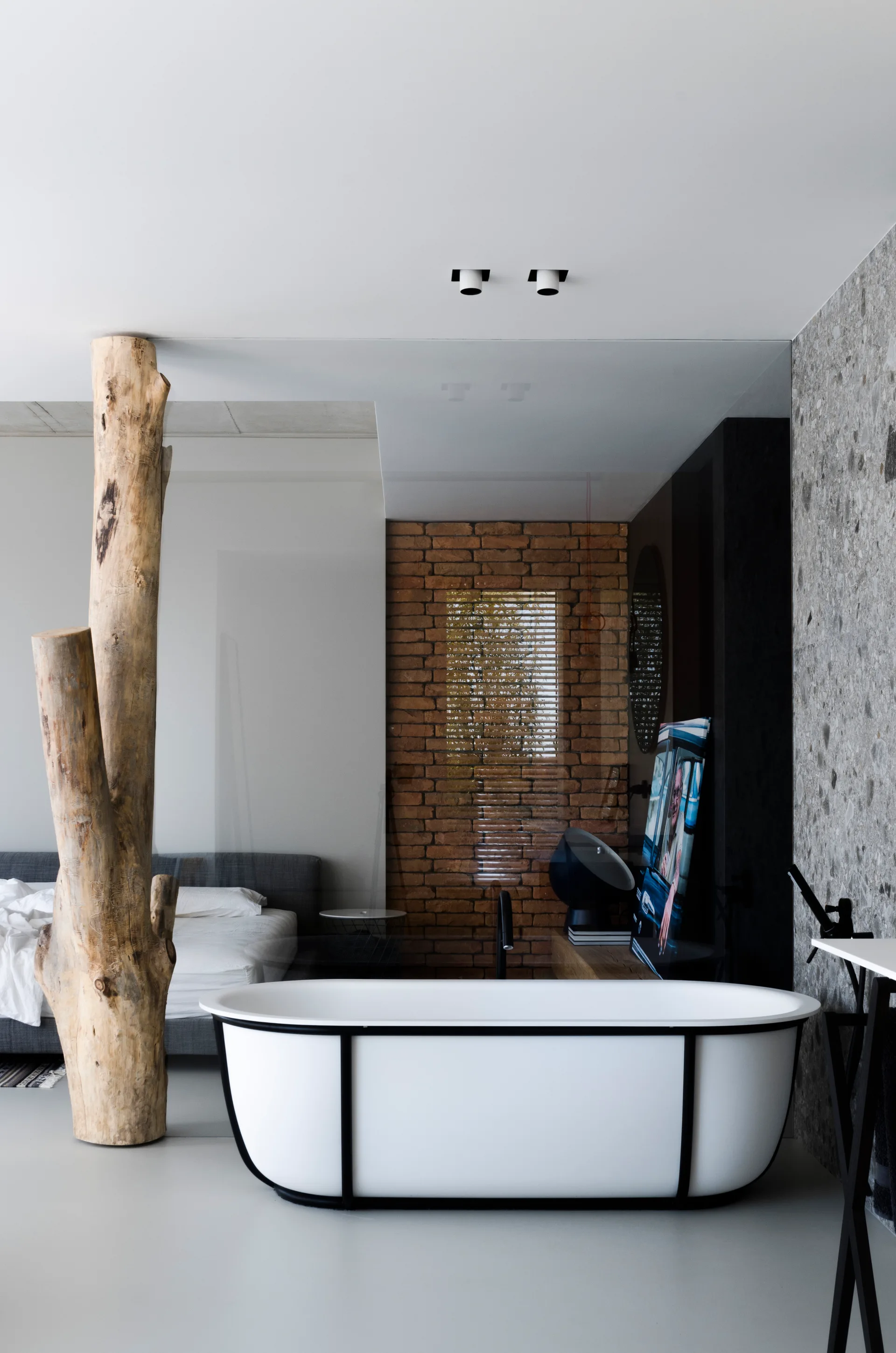 An open-plan bathroom showing a white freestanding tub with black metal detailing, a raw tree trunk pillar, and textured brick and stone walls.