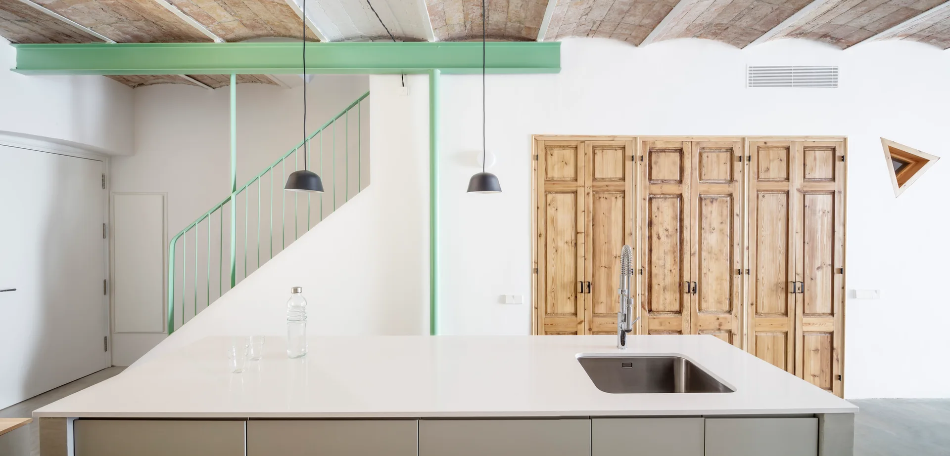 A modern kitchen with a white island, mint green ceiling beam, warm wood storage doors, and an exposed brick vaulted ceiling.