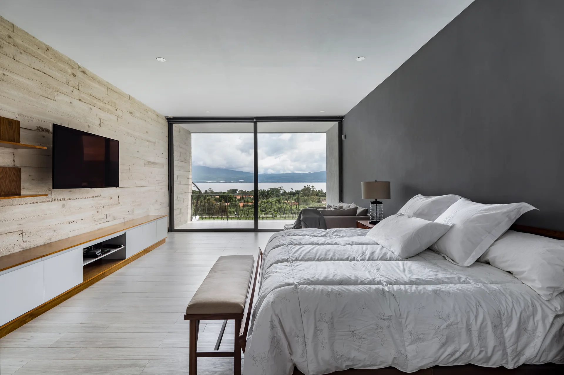 A contemporary bedroom features a bed with white bedding, a textured stone wall with a mounted television, and large sliding glass balcony doors.