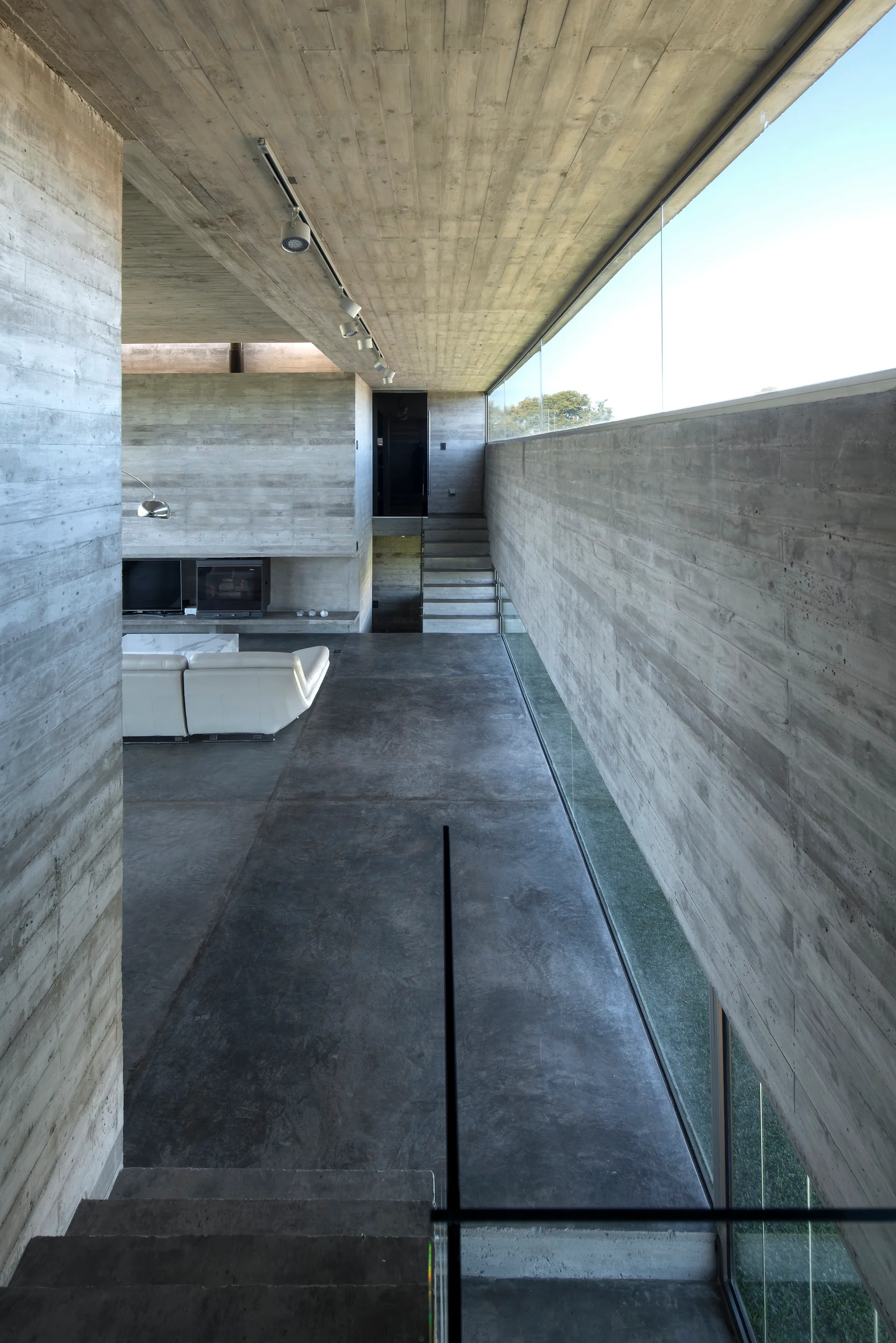 A minimalist living space with raw board-formed concrete walls, a dark concrete floor, a white sectional sofa, and a long clerestory window.