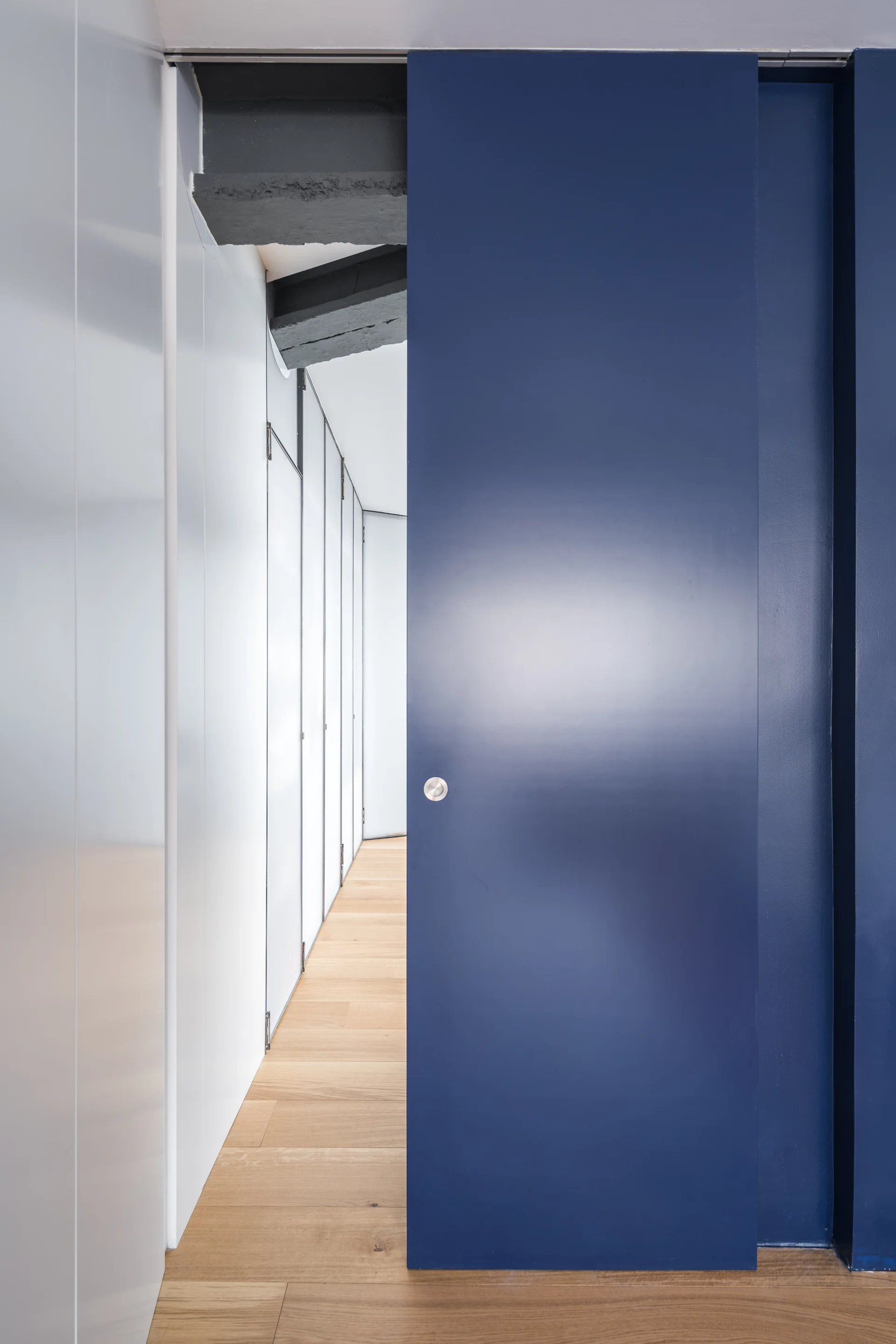A modern hallway features a large deep blue sliding door, white vertical wall panels, light wood flooring, and an exposed dark beam.