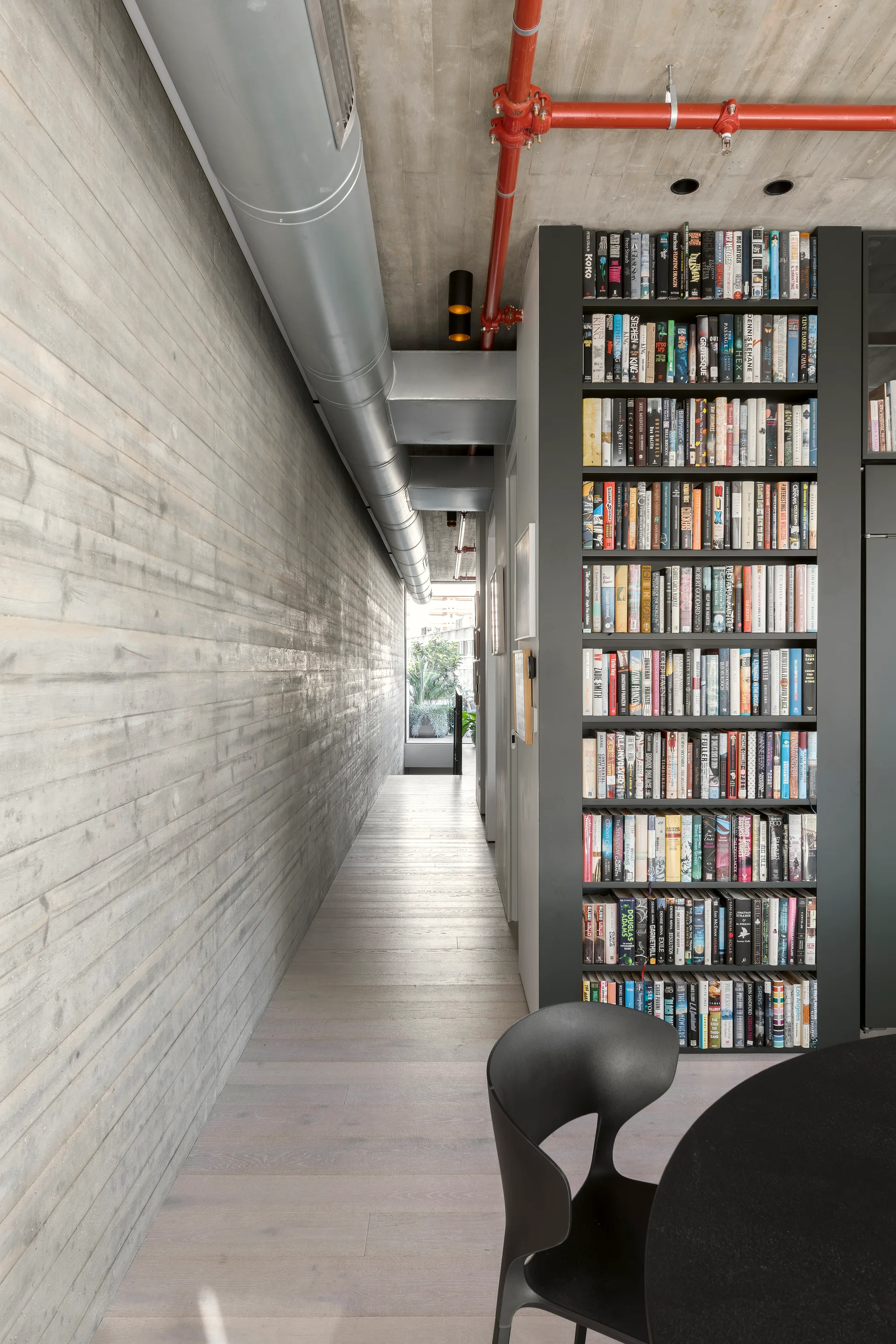 A narrow industrial hallway features a board-formed concrete wall, a tall black floor-to-ceiling bookshelf filled with books, and exposed ceiling pipes.