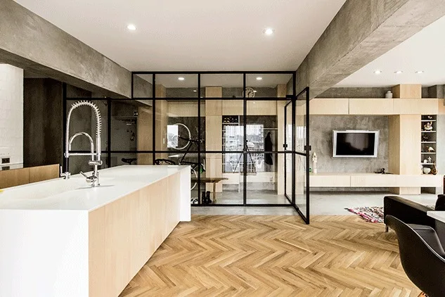 An open-plan apartment interior features a white kitchen island, herringbone wood flooring, and a black-framed glass partition separating the spaces.