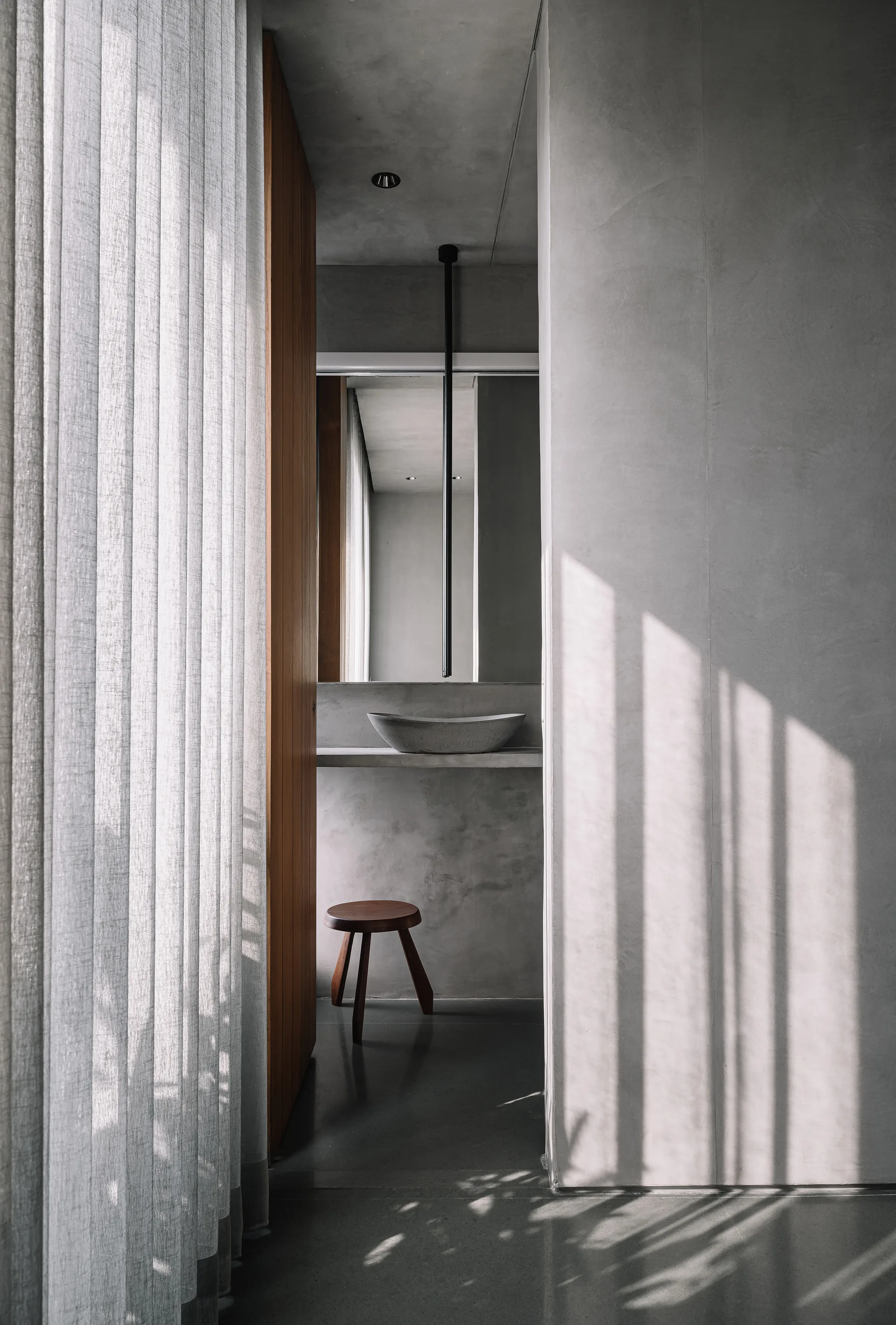 Minimalist concrete bathroom vanity with a stone basin, a ceiling-mounted faucet, a wooden stool, and sunlight filtering through a sheer curtain.