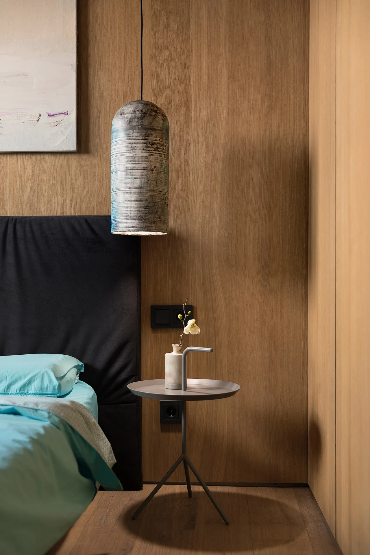 Modern bedroom corner with a black headboard, blue bedding, a small round side table, and a textured pendant lamp hanging against wood-paneled walls.