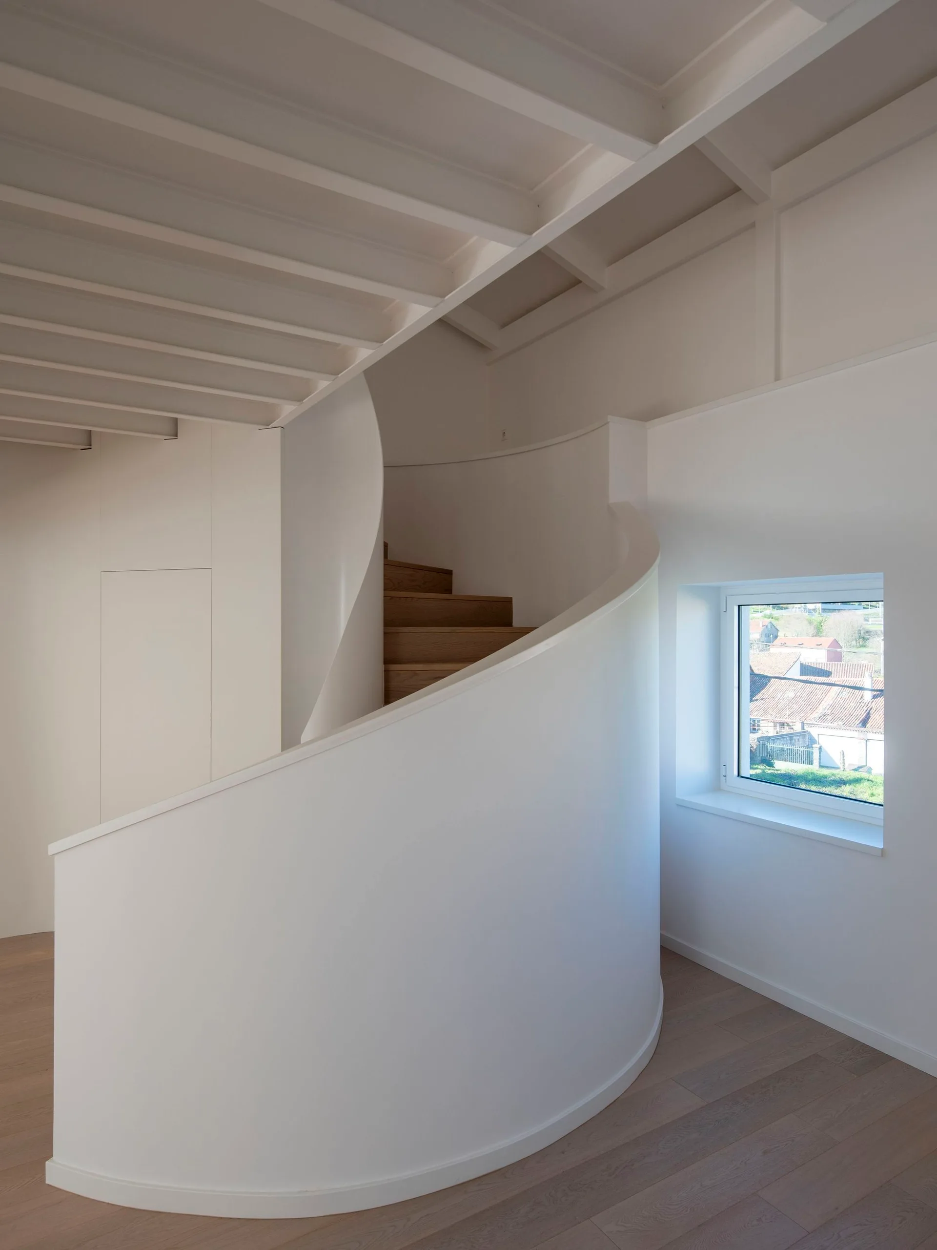 A minimalist white interior features a curved spiral staircase with light wood steps, exposed ceiling joists, and a small square window.