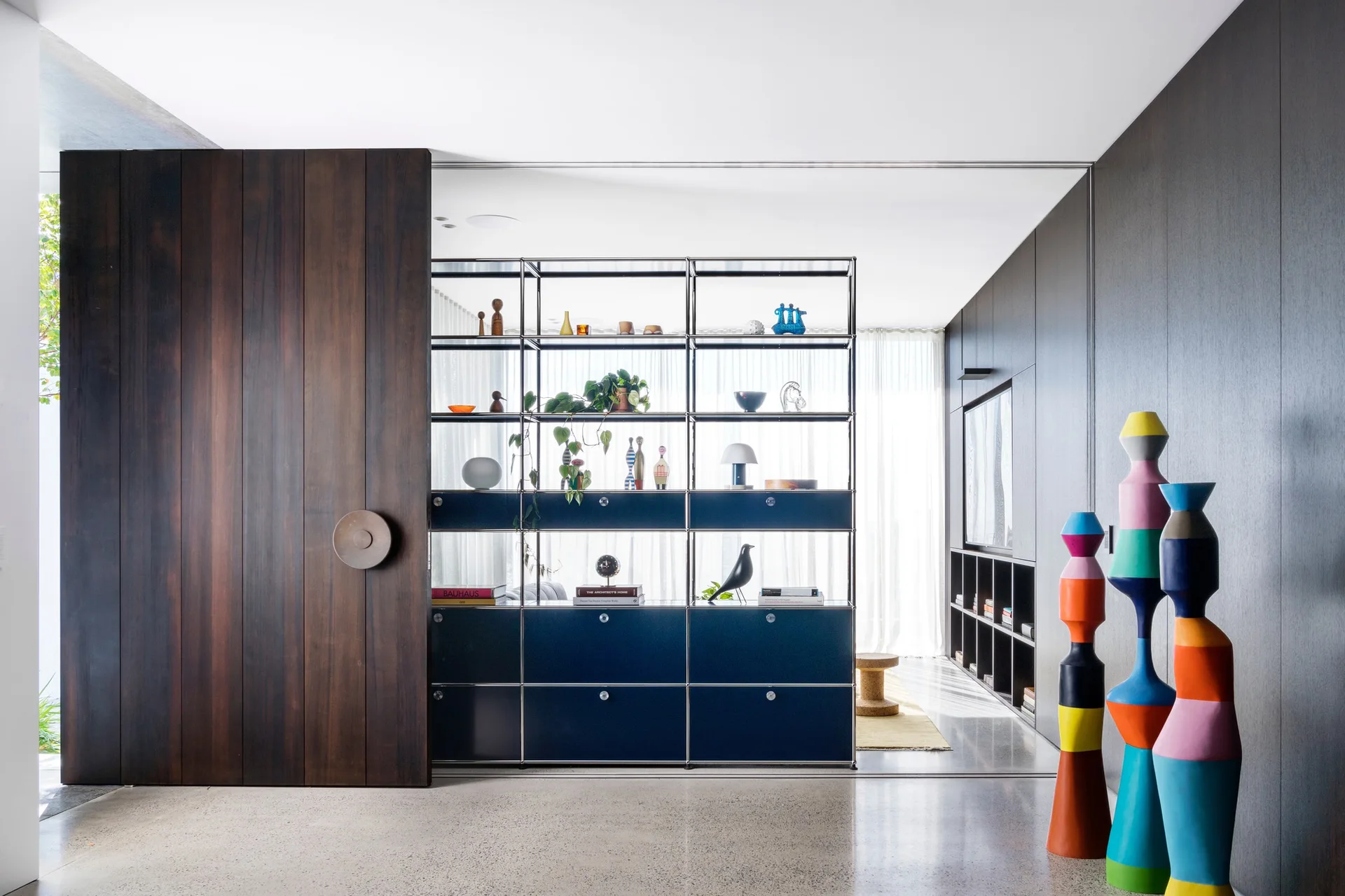 A modern interior with a dark wood sliding door, a black modular metal shelving unit, and colorful stacked sculptures on a concrete floor.