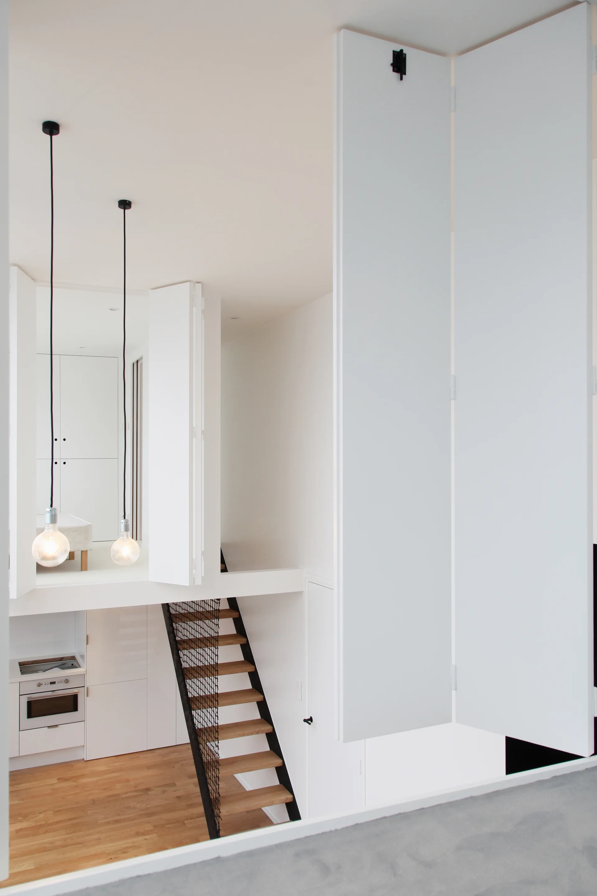 A modern split-level loft featuring a wooden staircase with black wire mesh railing, bare bulb pendant lights, and tall white folding doors.