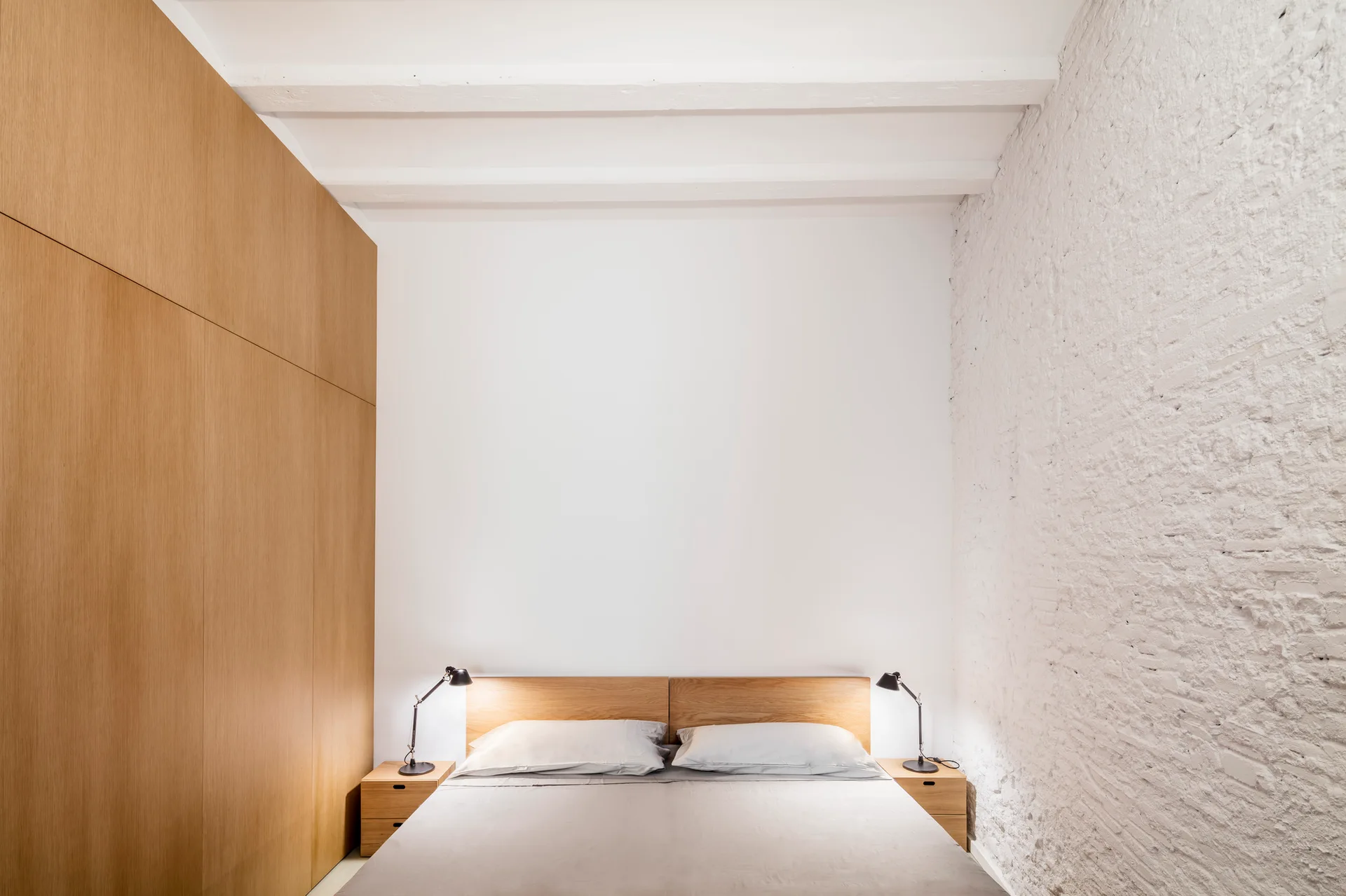 A minimalist bedroom features a bed with a wooden headboard flanked by integrated wood paneling on one side and a textured white brick wall on the other.