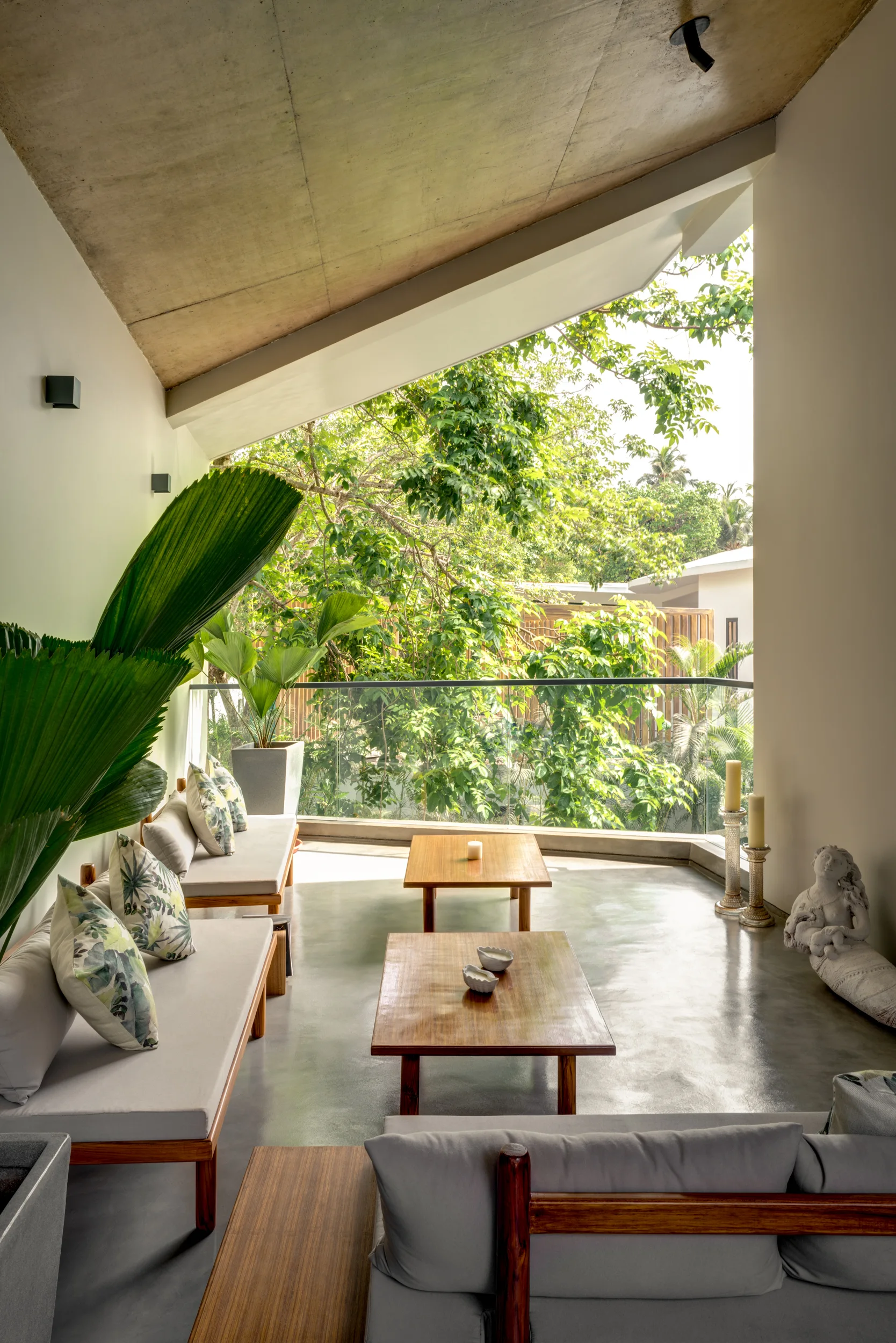An open-air patio features grey concrete surfaces, low-profile wood sofas with patterned pillows, and wooden coffee tables overlooking a green garden.