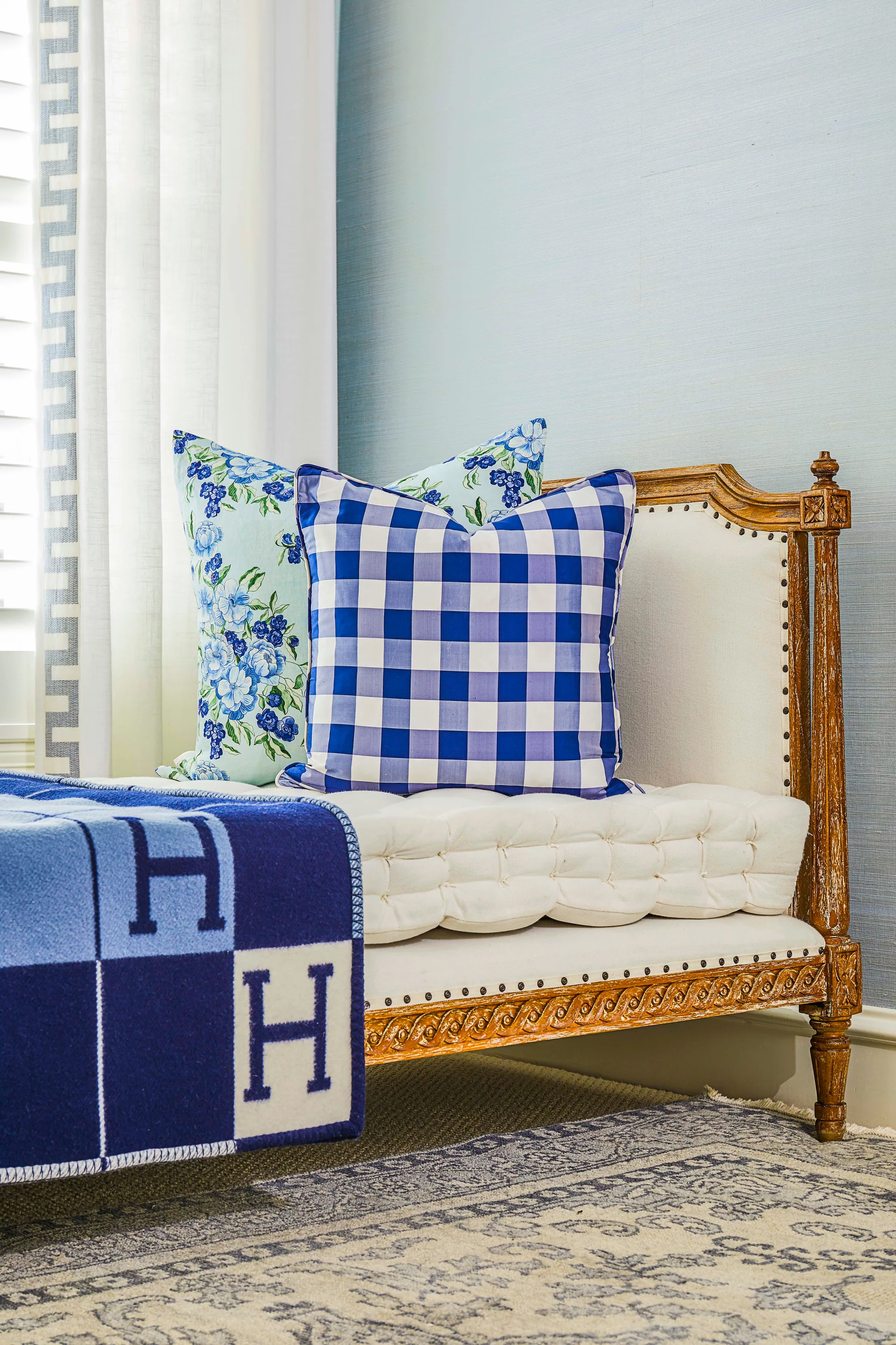 A cream tufted wooden bench with blue gingham and floral pillows, draped with a blue monogrammed blanket in front of a blue wall.
