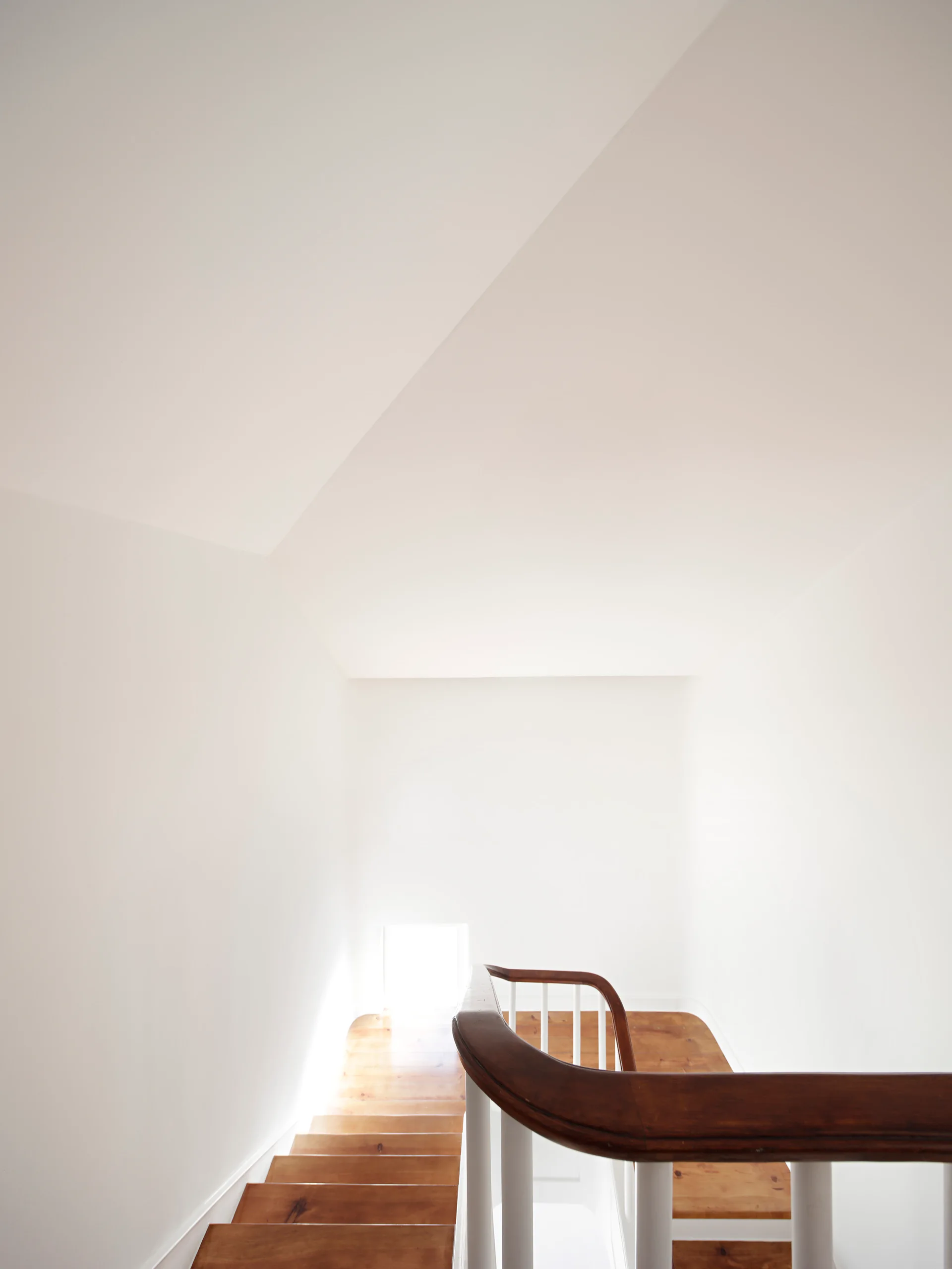 Looking down a minimalist white stairwell with varnished wood steps and a curved dark brown handrail.