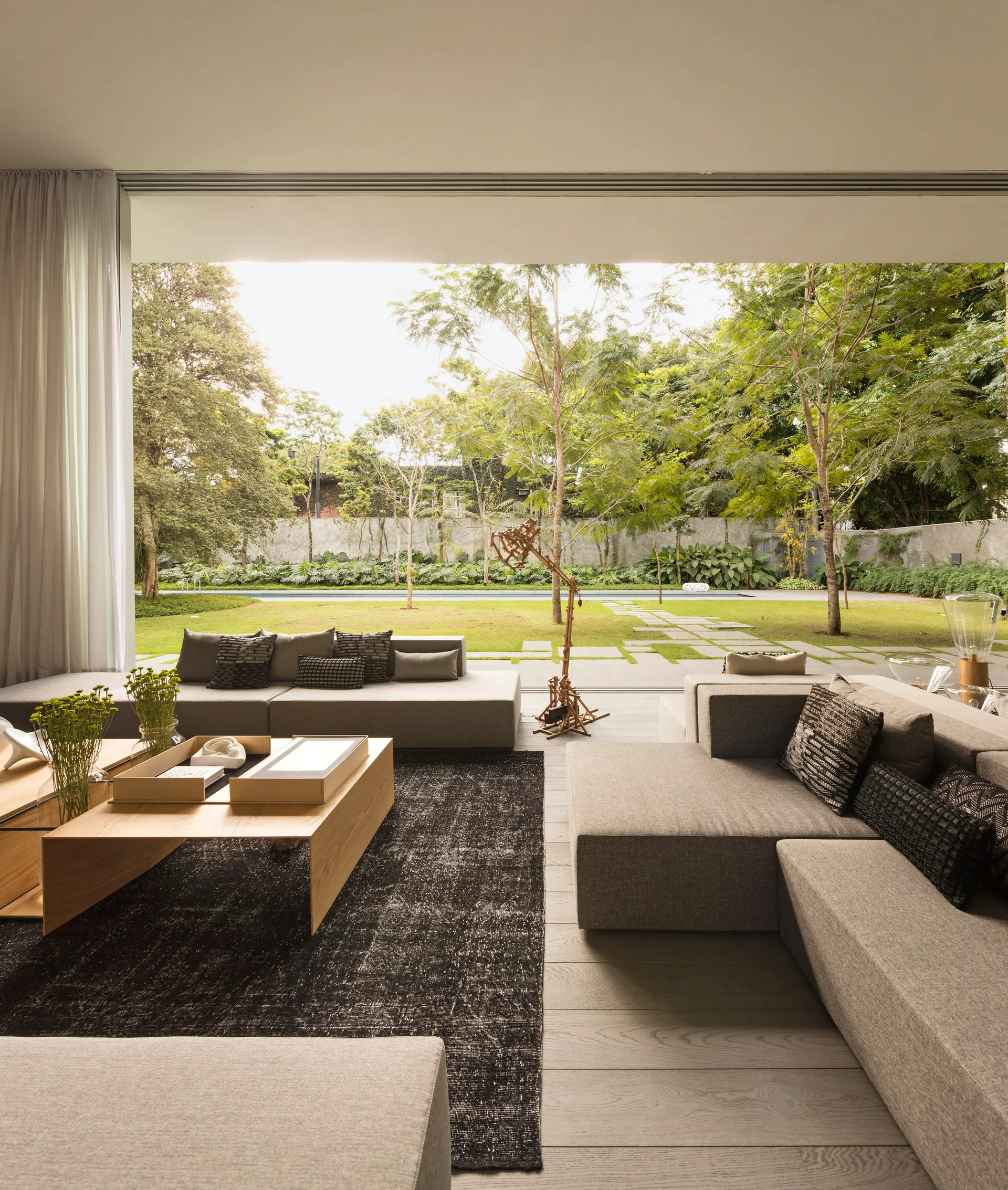 A contemporary living room with gray modular sofas and low wood coffee tables faces a large sliding glass doorway opening to a green garden.