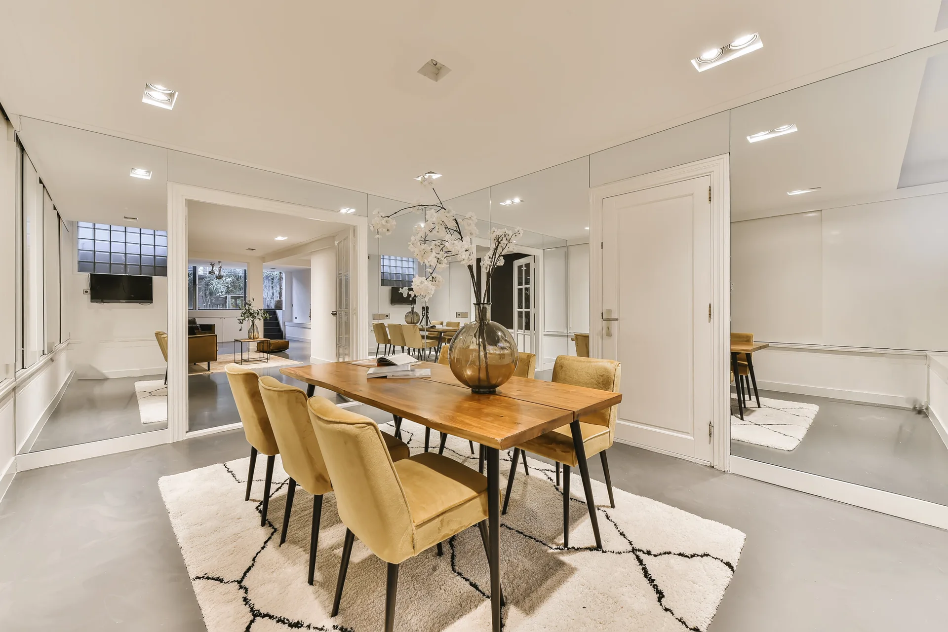 A wooden dining table surrounded by yellow upholstered chairs sits on a patterned shag rug next to a full-height mirrored wall.
