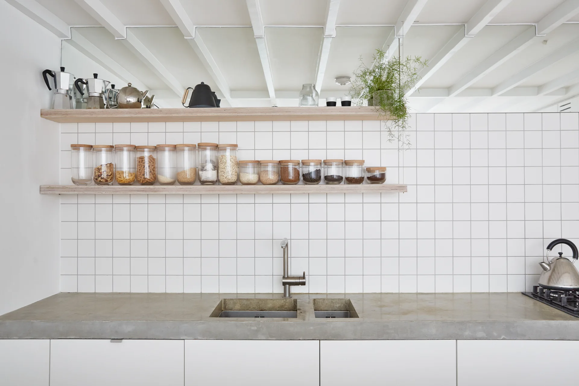 A kitchen features a white grid tile backsplash, two open wooden shelves lined with glass jars, and an integrated double sink in a concrete countertop.