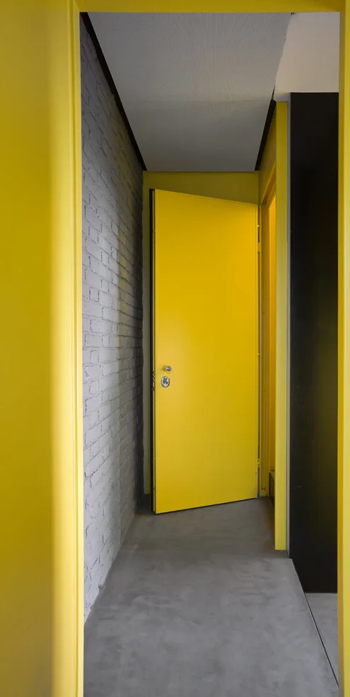 A modern hallway features a bright yellow door, a grey painted brick wall, and a smooth concrete floor under natural light.