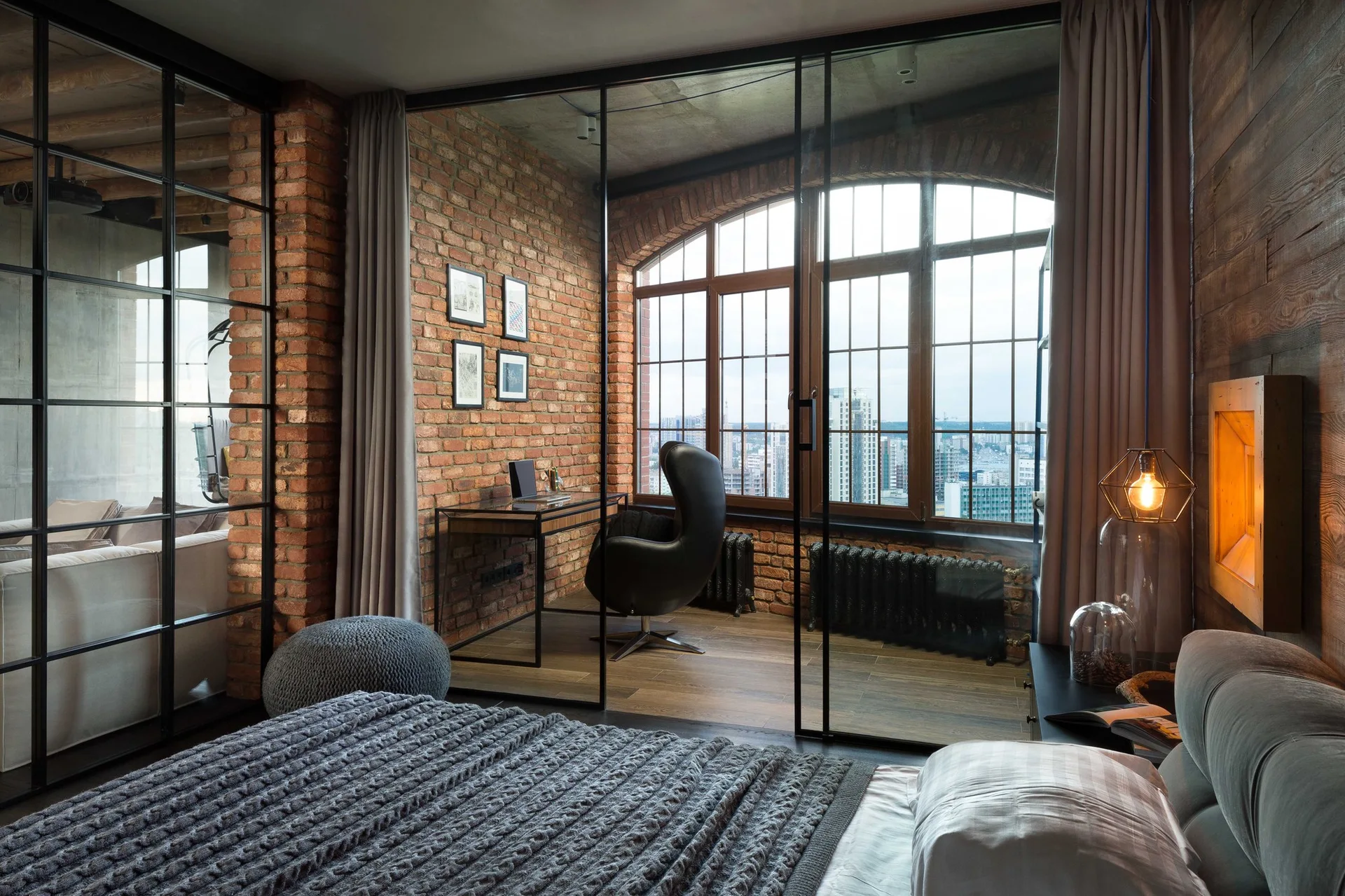 A modern industrial bedroom features a black metal-framed glass partition separating the grey bed from a brick-walled home office nook.