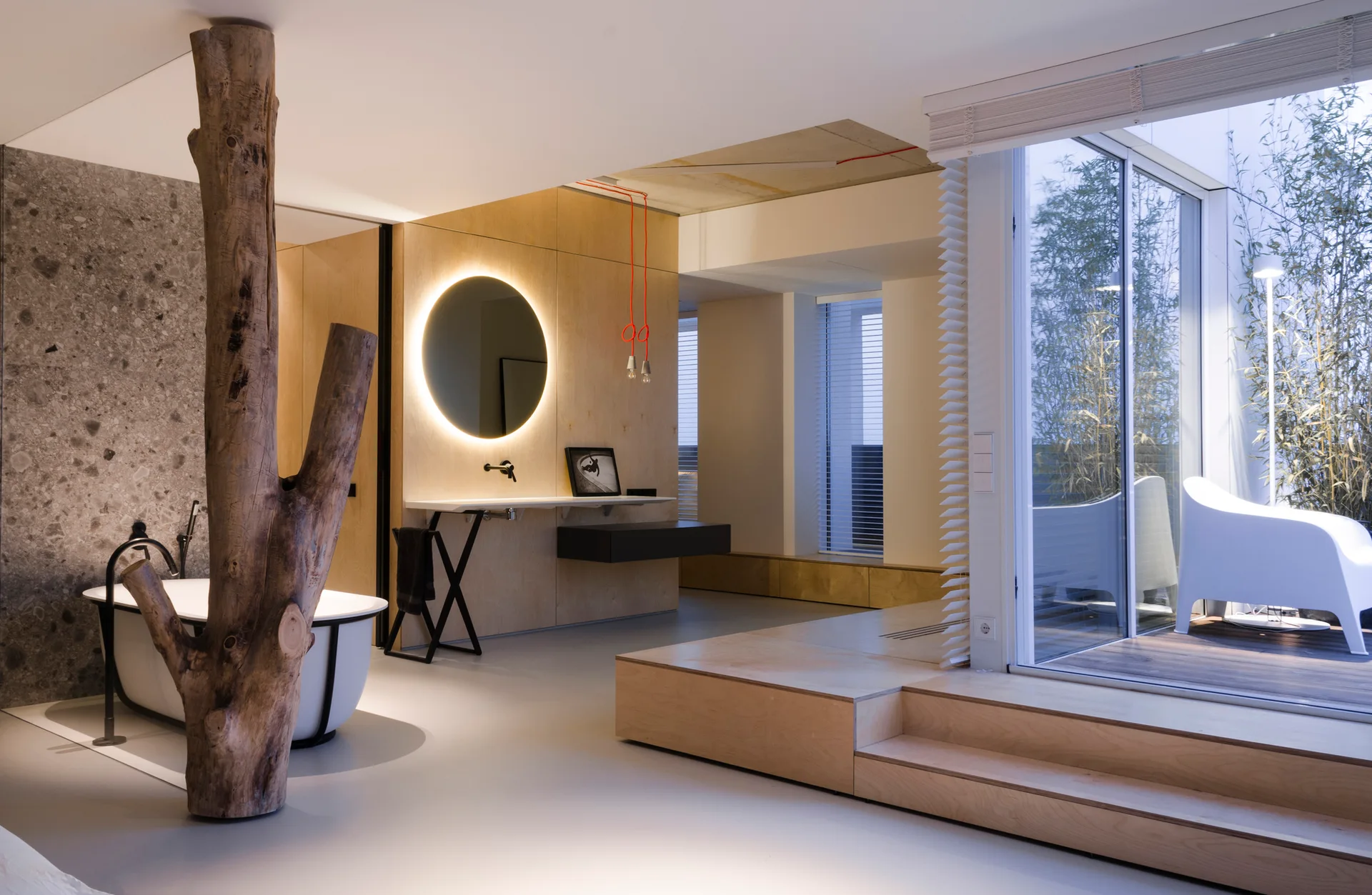 A modern bathroom with a freestanding bathtub next to a large tree trunk column, a backlit round mirror on a plywood wall, and steps to a terrace.