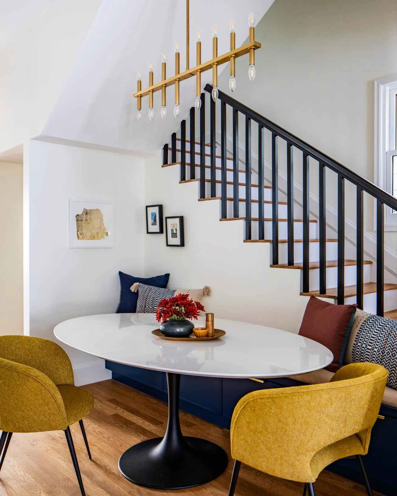 A compact dining nook features a white oval pedestal table, two yellow armchairs, and a navy blue banquette built under a wooden staircase.
