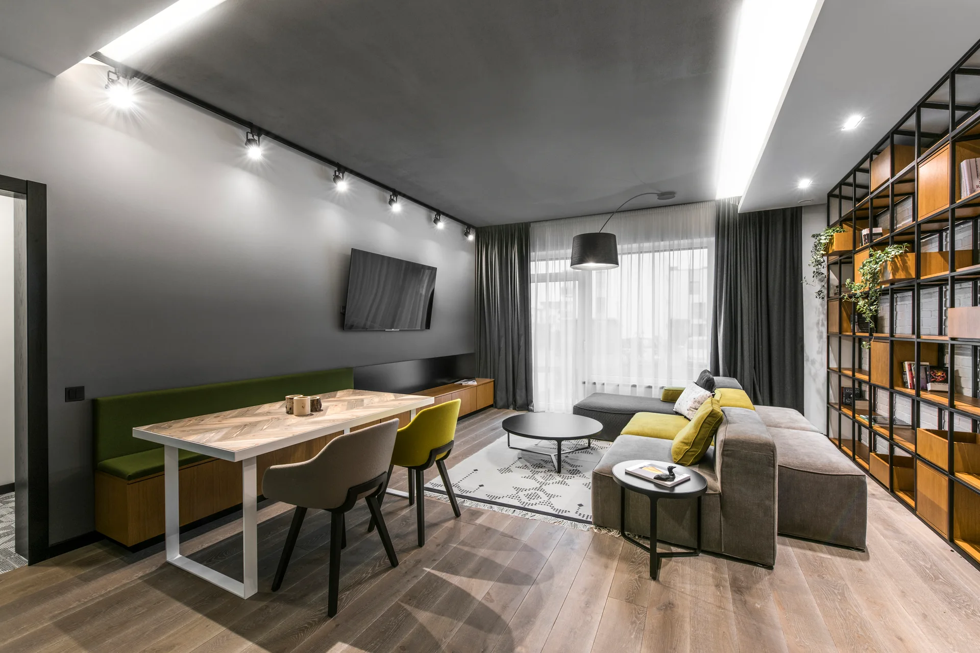 A modern gray living room features a modular sectional sofa, a chevron-patterned dining table with a built-in bench, and a tall metal bookshelf.