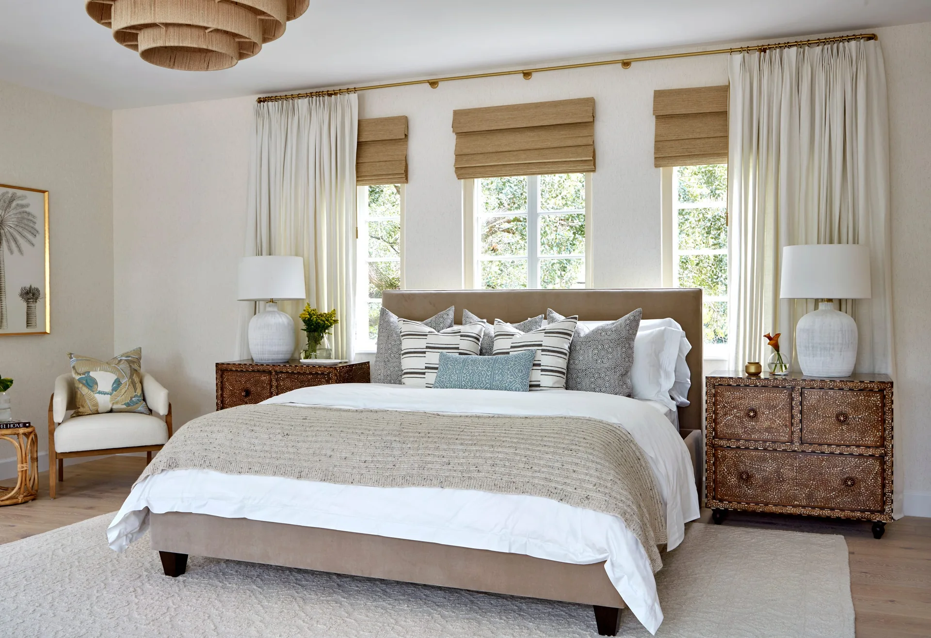 A bedroom features an upholstered beige bed flanked by patterned wooden nightstands and illuminated by white lamps under three windows.