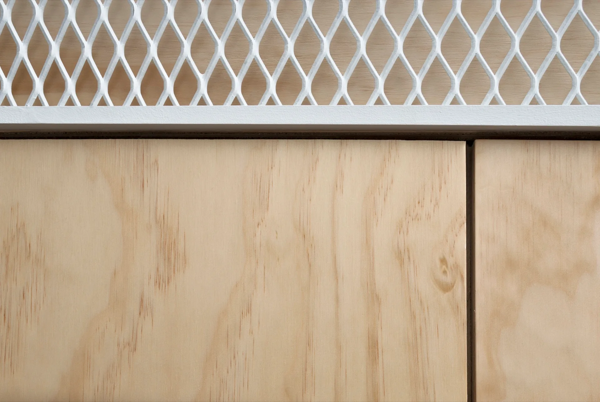 A close-up view of a light-colored natural wood cabinet panel situated below a white painted metal mesh screen with a diamond pattern.