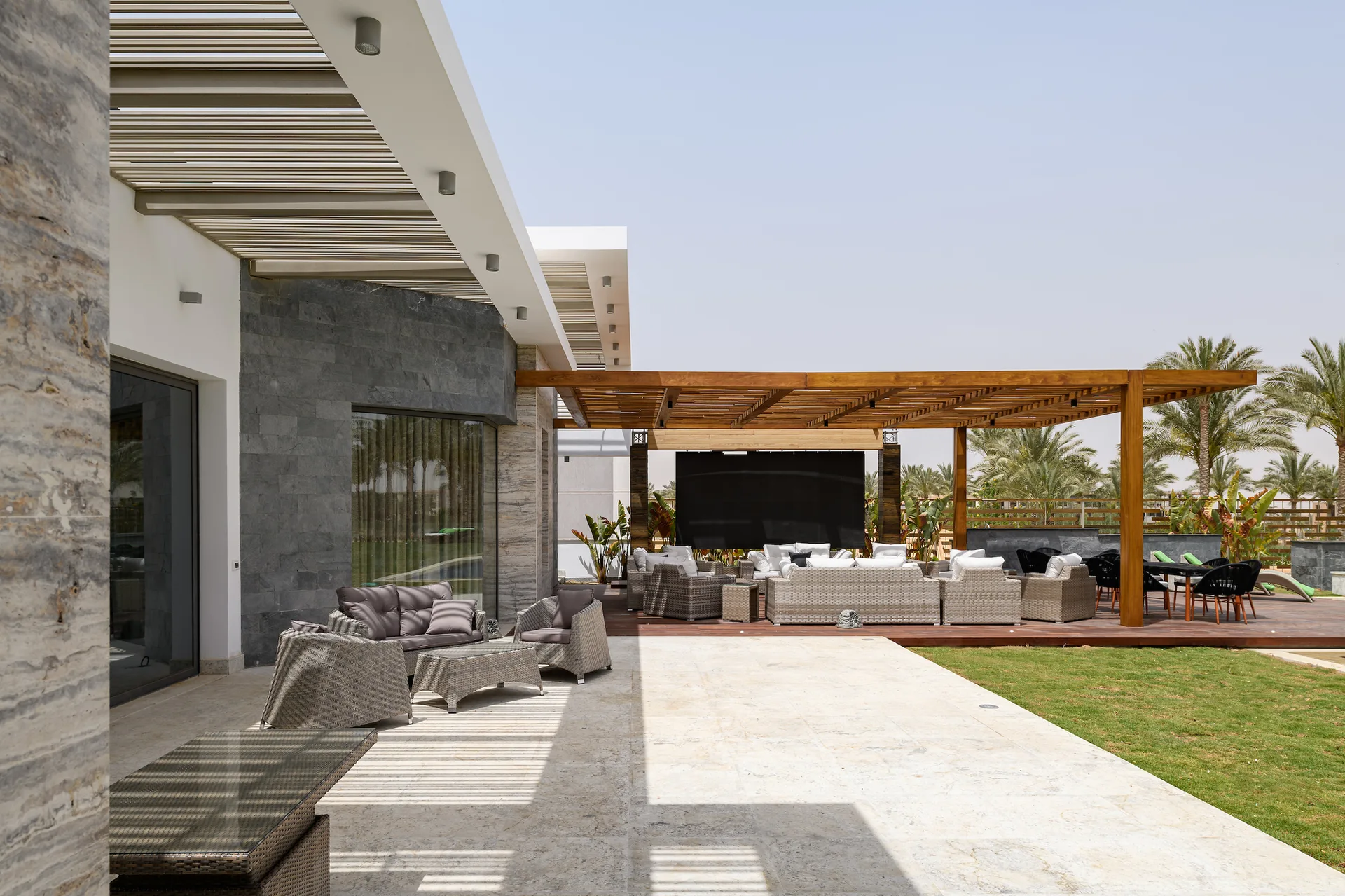 A modern outdoor patio featuring grey wicker furniture on a stone terrace and a raised wooden deck covered by a wood slat pergola next to a lawn.