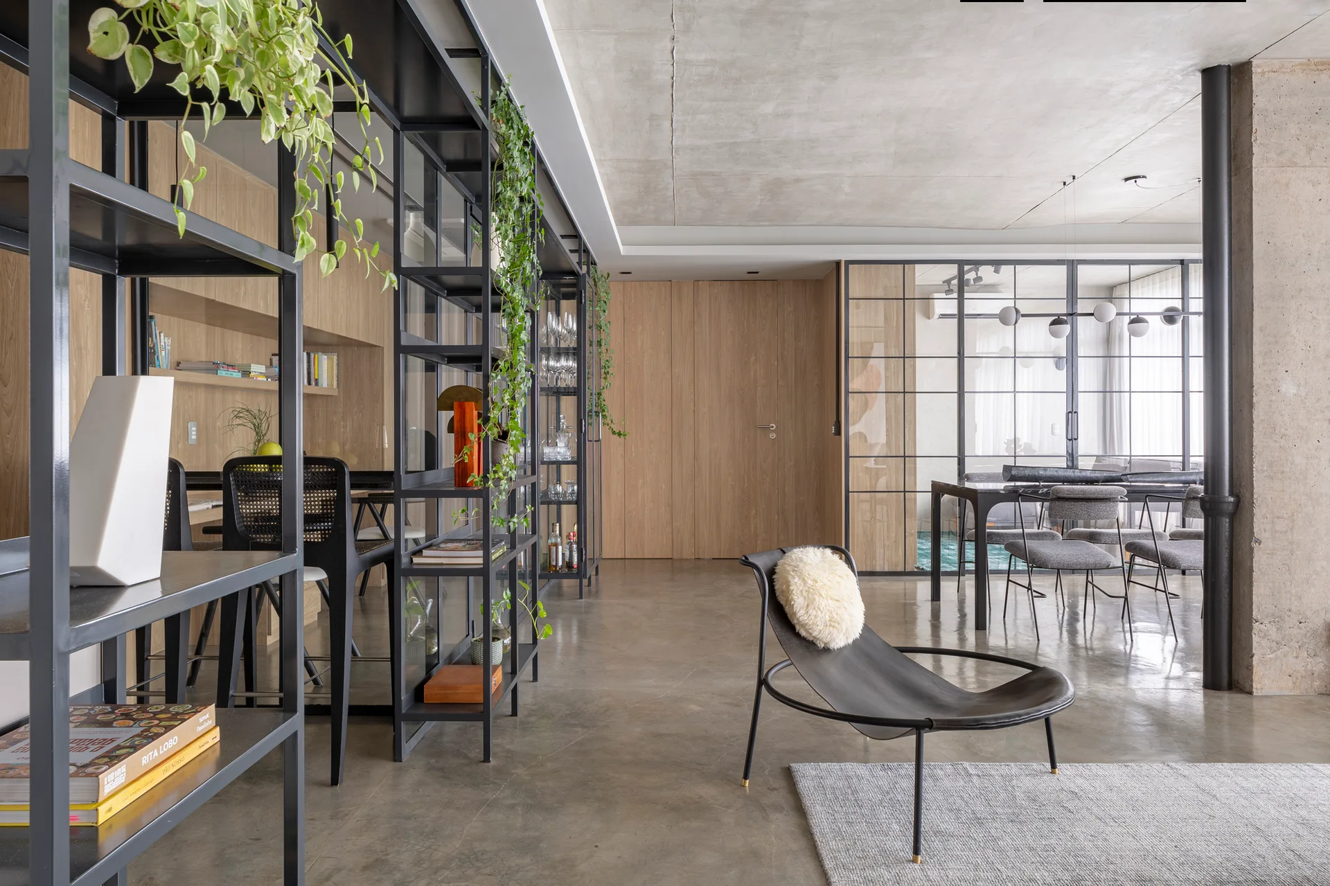 A contemporary open-plan living room featuring a black metal shelving unit, a curved black accent chair, polished concrete floors, and a concrete ceiling.