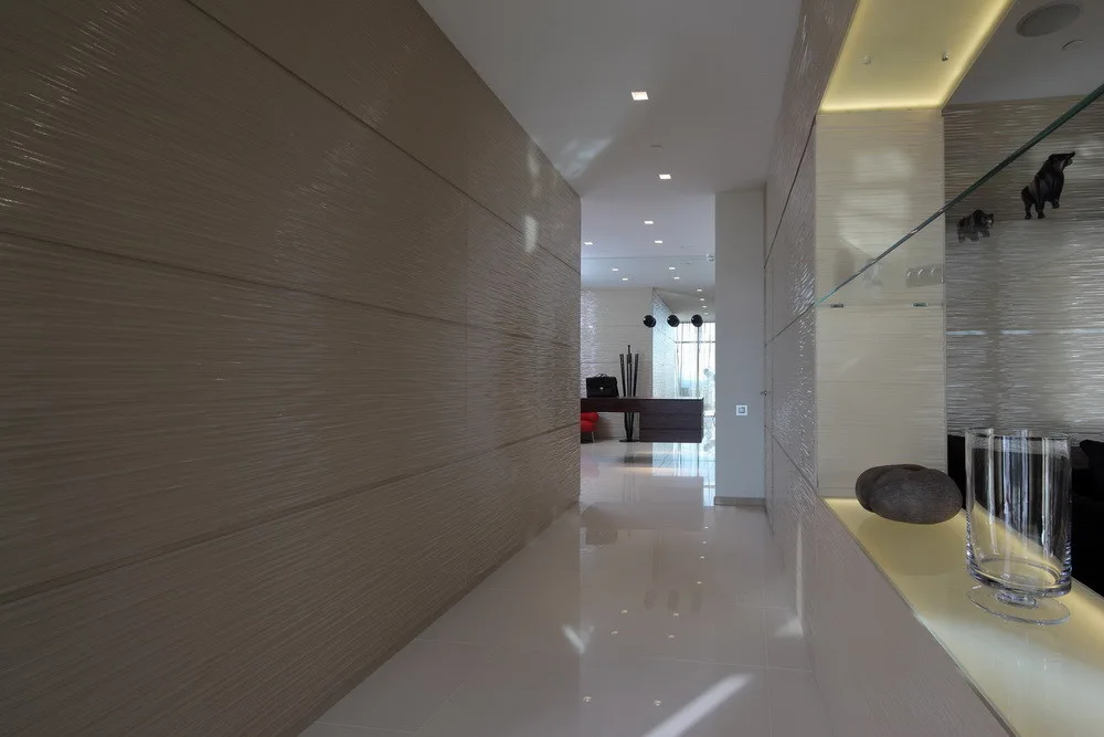 A modern hallway features textured beige wall panels, a highly reflective white tile floor, and an illuminated display niche.