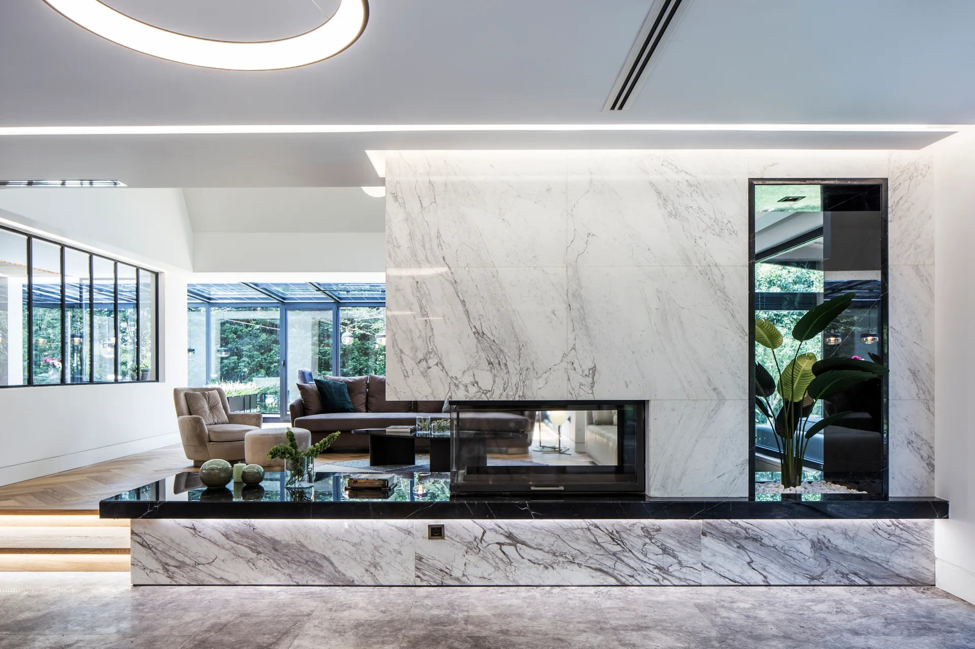 A modern living room features a large veined marble fireplace divider with a black stone hearth, recessed LED lighting, and a plant niche.