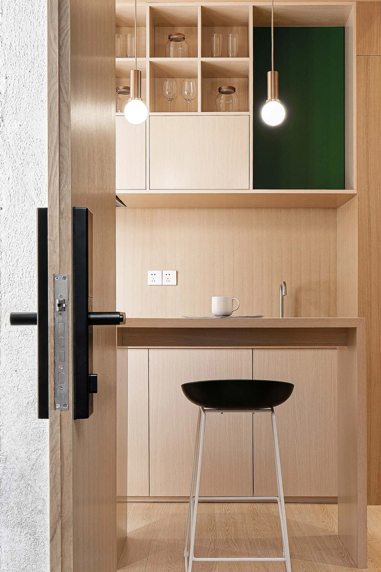 A view through an open wooden door reveals a minimalist wood kitchenette with open shelving, a green accent panel, and two hanging globe lights.