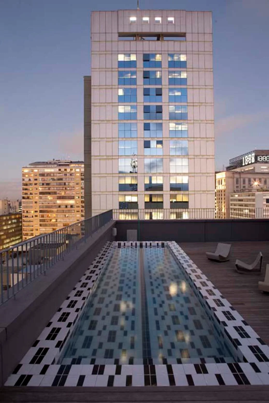 An elongated rooftop swimming pool featuring a black and white geometric tile pattern on a wood deck overlooking a city skyline at twilight.