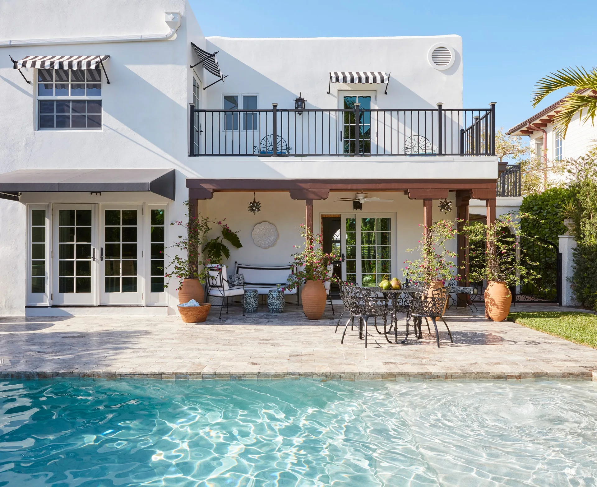 A white stucco house features a stone paver patio with wrought iron furniture, large terracotta planters, and a blue swimming pool.