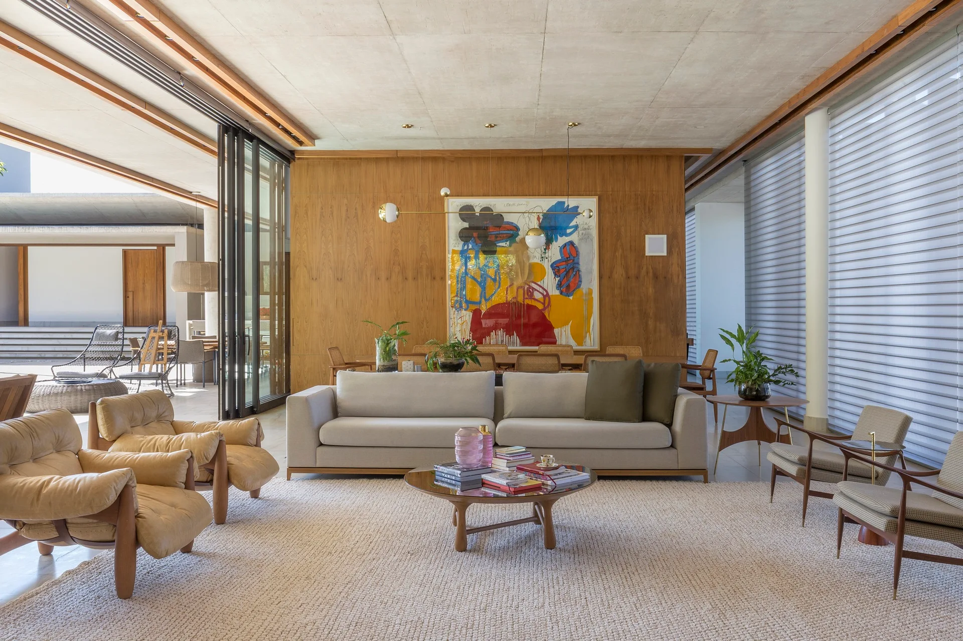 A spacious living room features a beige sofa, tan leather chairs, and a round coffee table against a wood-paneled wall under a concrete ceiling.