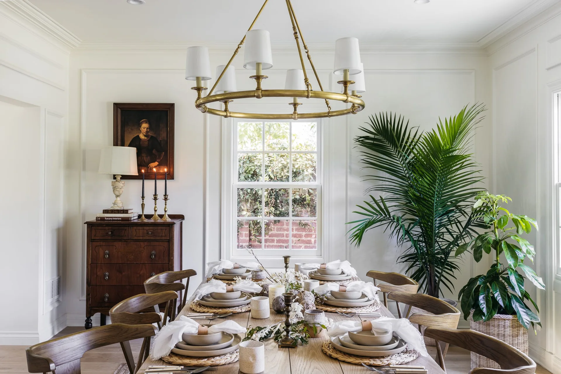 A set wooden dining table sits under a brass chandelier in a white dining room with panel molding and a dark antique chest.